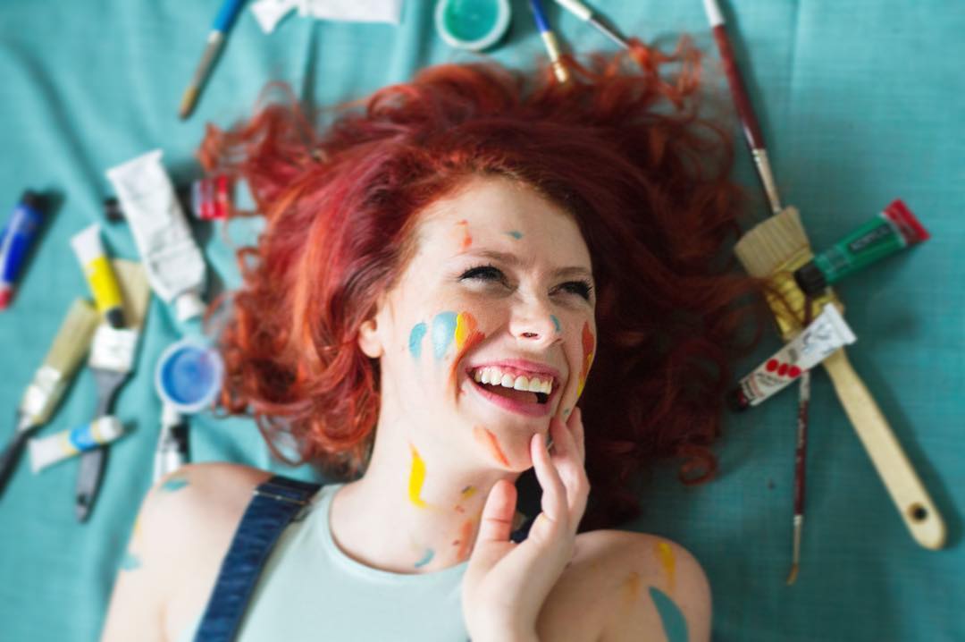Smiling woman with red hair lying on the floor, her face and arms covered in colorful paint, surrounded by brushes and paint tubes.
