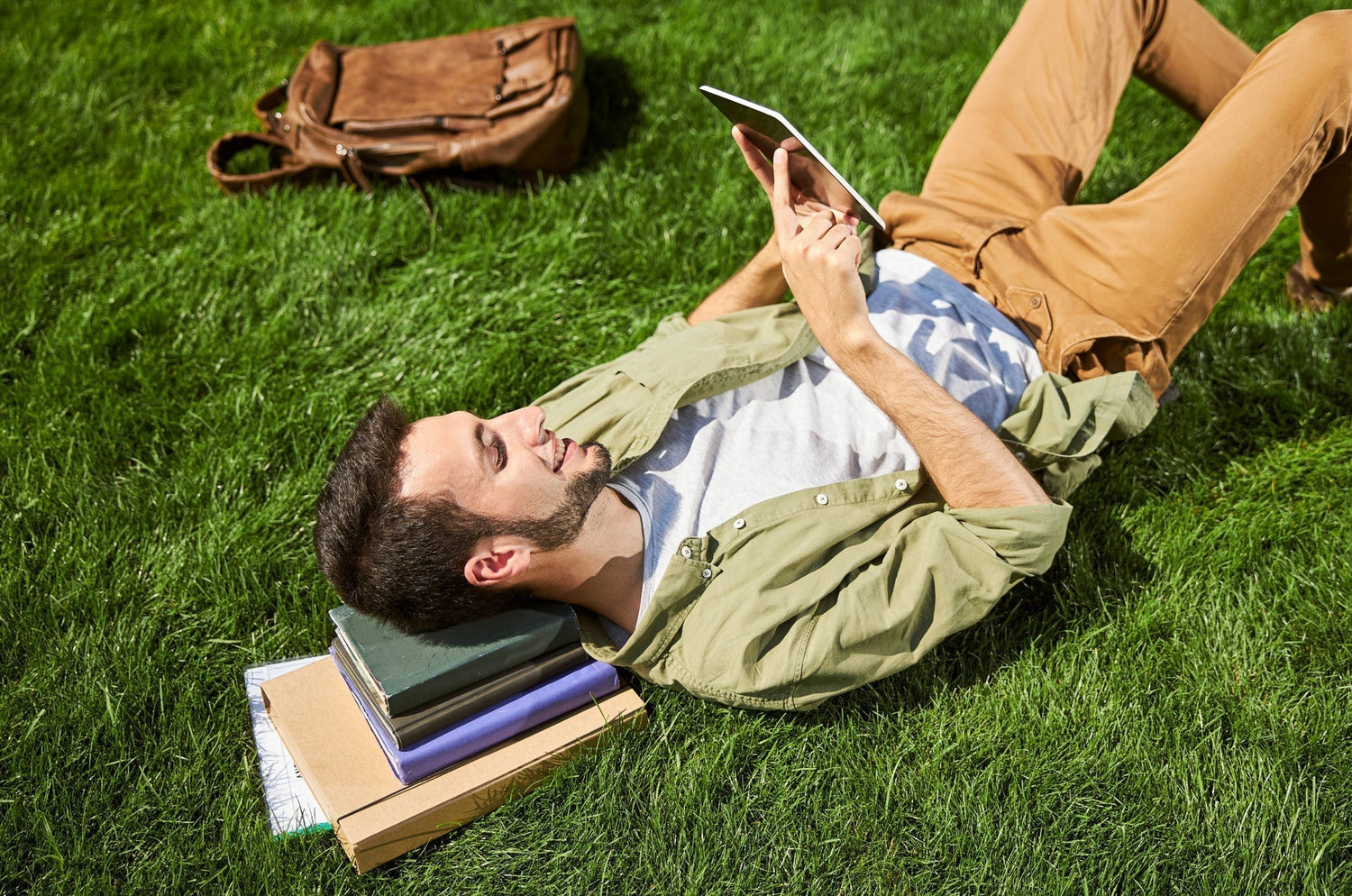 Man lying on the grass outdoors, smiling while using a tablet. He rests his head on a stack of books with a backpack nearby.