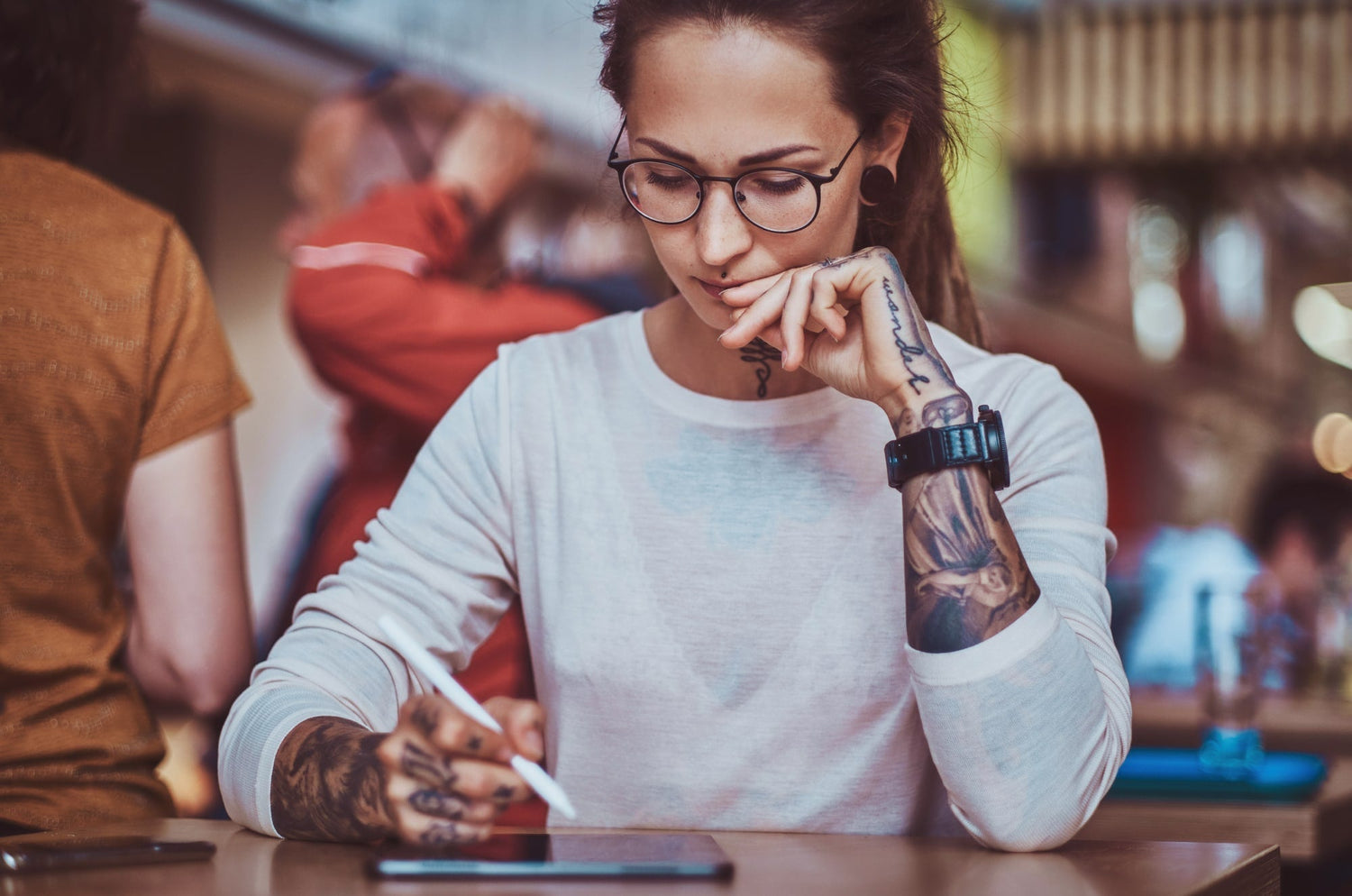 Person with glasses and tattooed arms sitting at a table, focused on writing or drawing on an iPad with an Apple Pencil in a busy café setting.