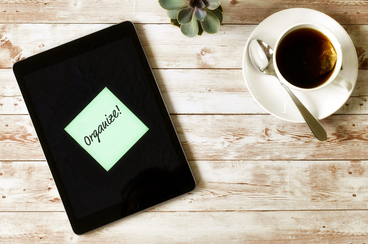 Flat lay of an iPad on a wooden table displaying a digital sticky note with the word ‘Organize!’ next to a cup of tea and a succulent plant.