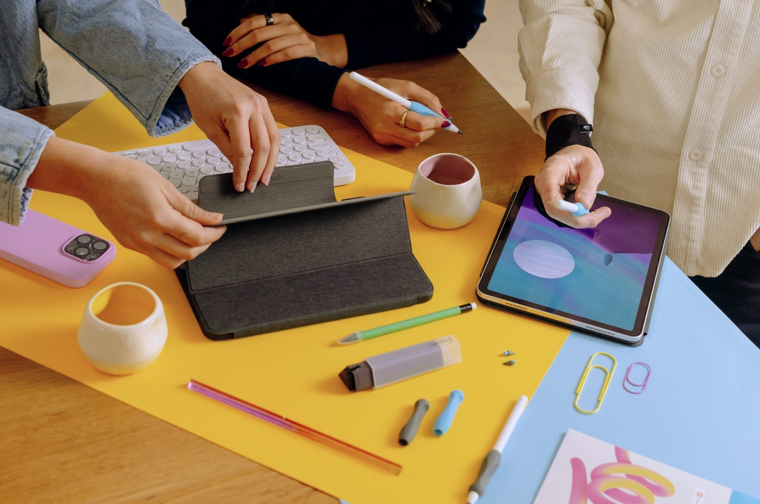 Creative workspace with people using iPads, Apple Pencils, and Paperlike™ accessories on a colorful desk covered with stationery, keyboard, and tablet cases.