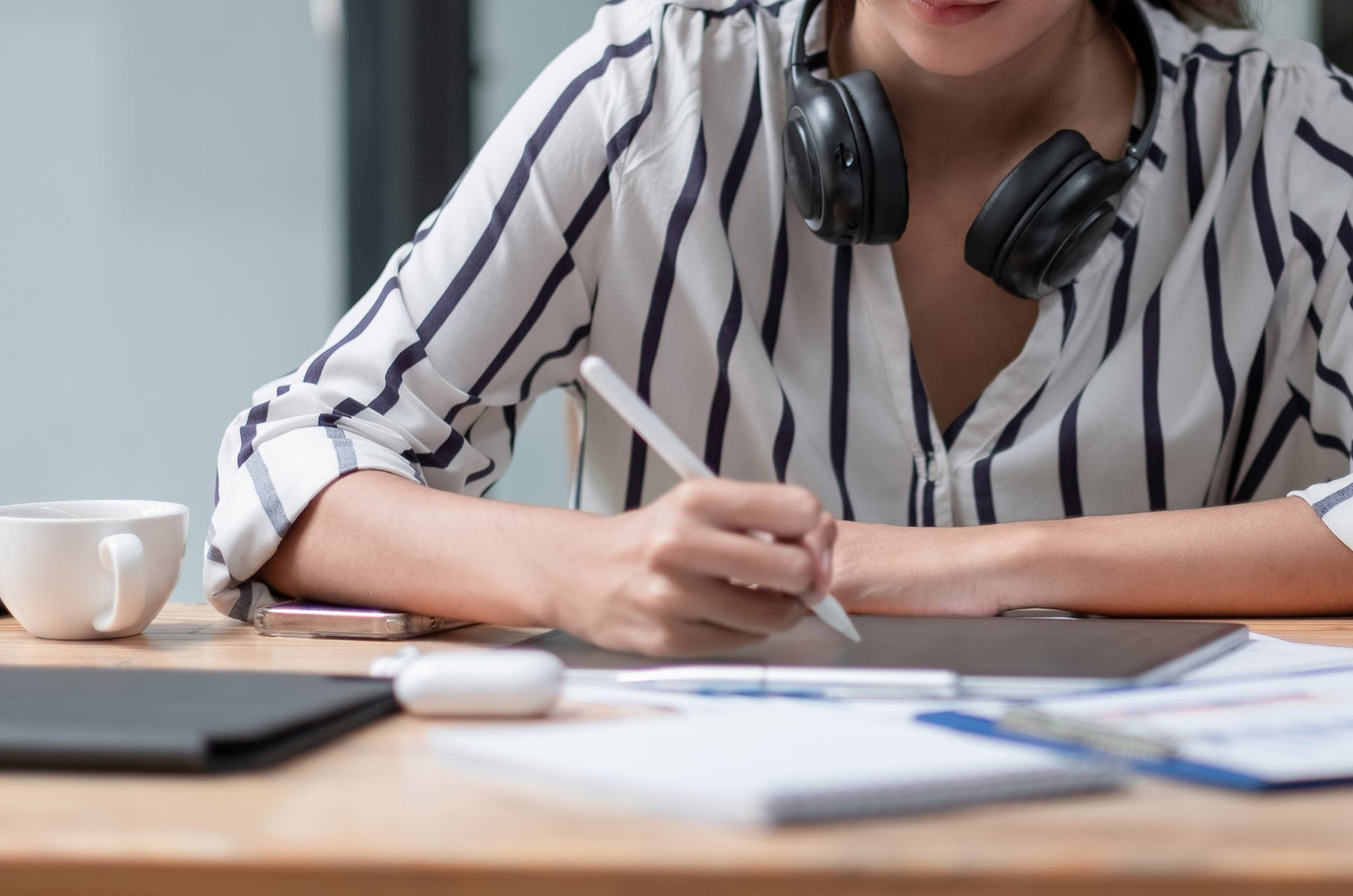 Person in a striped shirt with headphones around their neck using an Apple Pencil on an iPad at a desk with notebooks, papers, and a coffee cup.