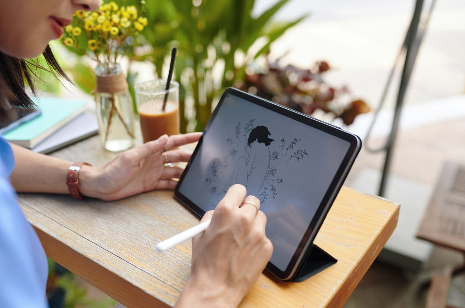Digital artist sketching floral line art on iPad with Apple Pencil at a coffee shop.