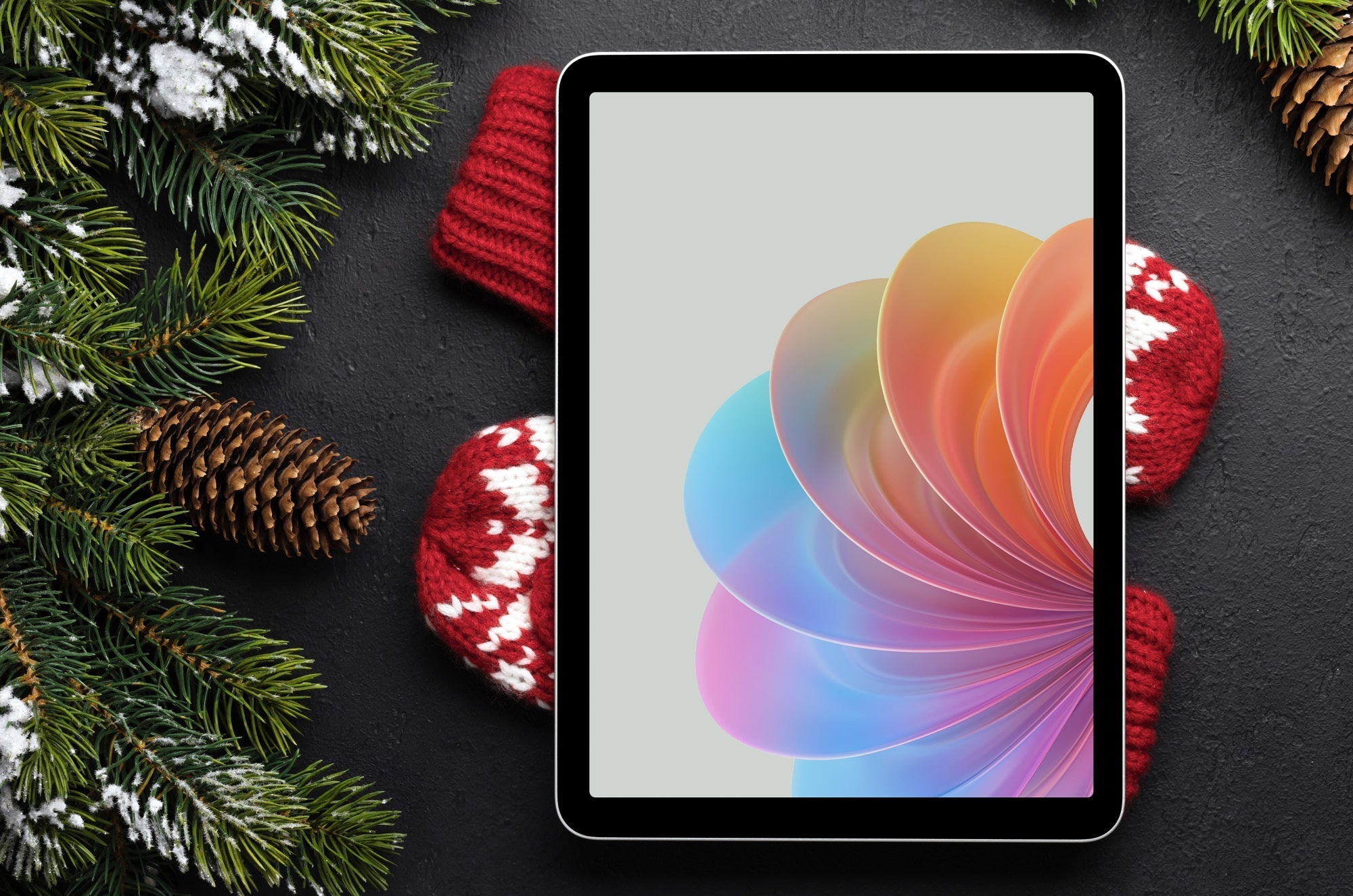 An iPad with colorful swirls on the screen, lying on a pair of red and white mittens and surrounded by pinecones and tree branches.