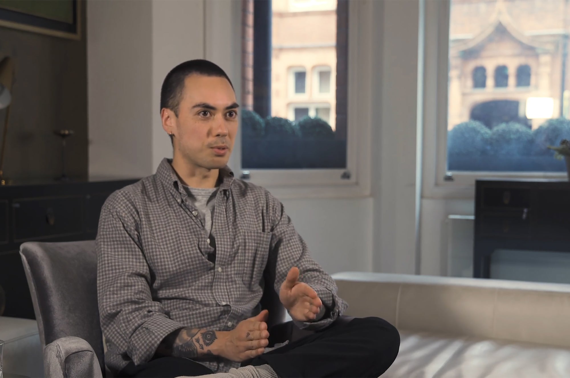 Person sitting cross-legged on a couch in a bright room, wearing a checkered shirt, speaking with hands gesturing during an interview-style conversation.