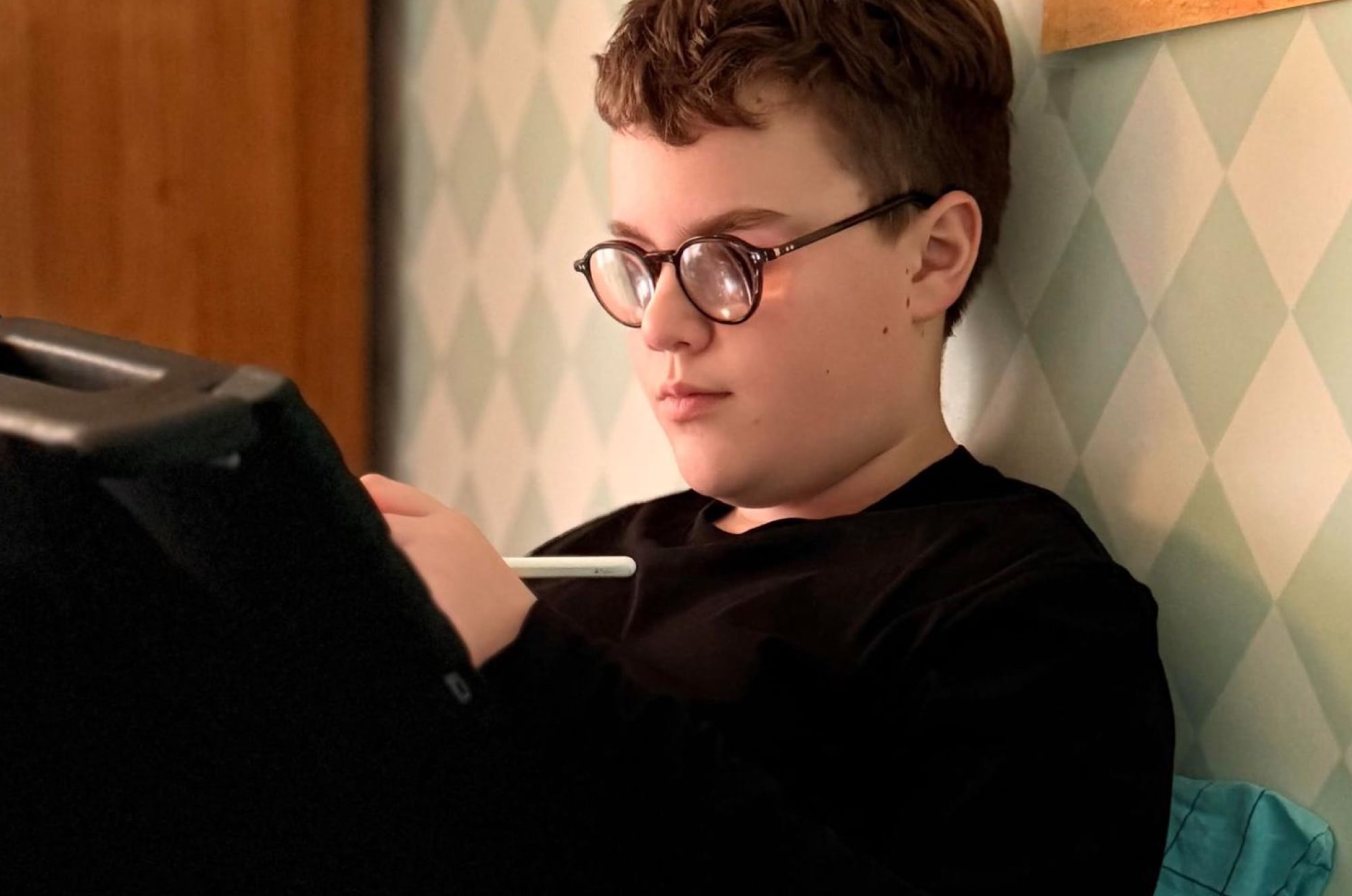 A boy sitting on a bed, sketching on an iPad with an Apple Pencil.