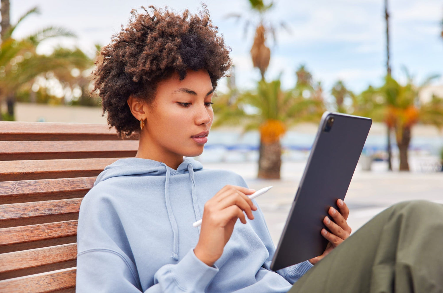 Person in a light blue hoodie sitting on a wooden bench outdoors, holding an iPad and Apple Pencil with palm trees in the background.