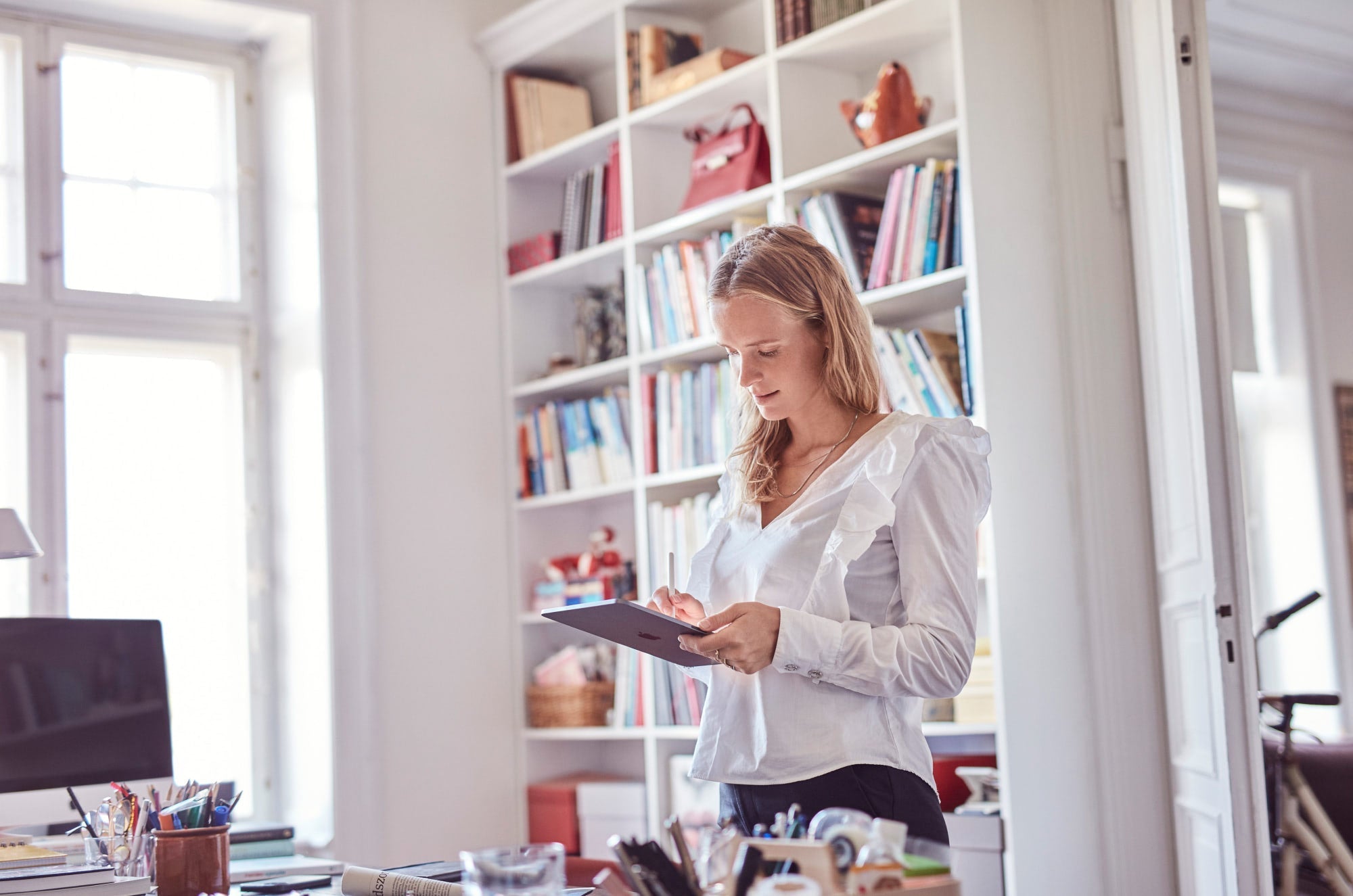 Woman using iPad with Apple Pencil in a bright home office with bookshelf in the background.