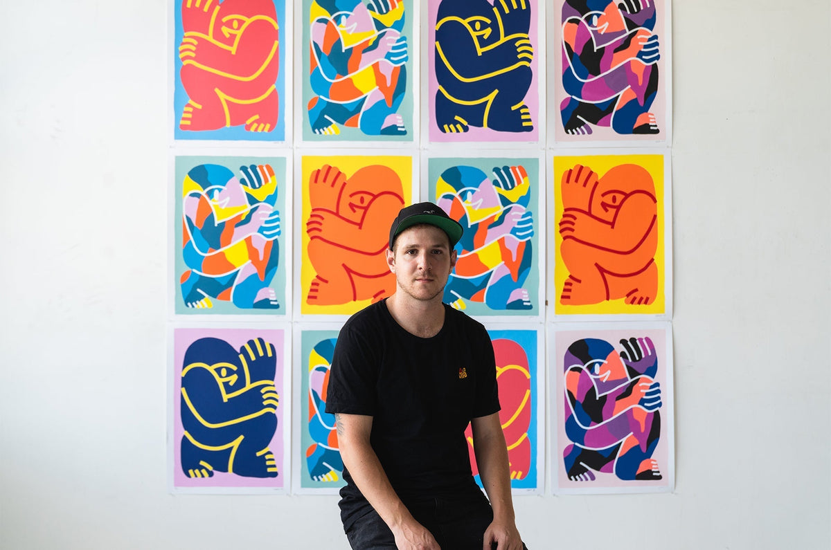 Man in black t-shirt and cap sitting in front of a wall filled with colorful abstract artwork featuring bold, stylized human figures.