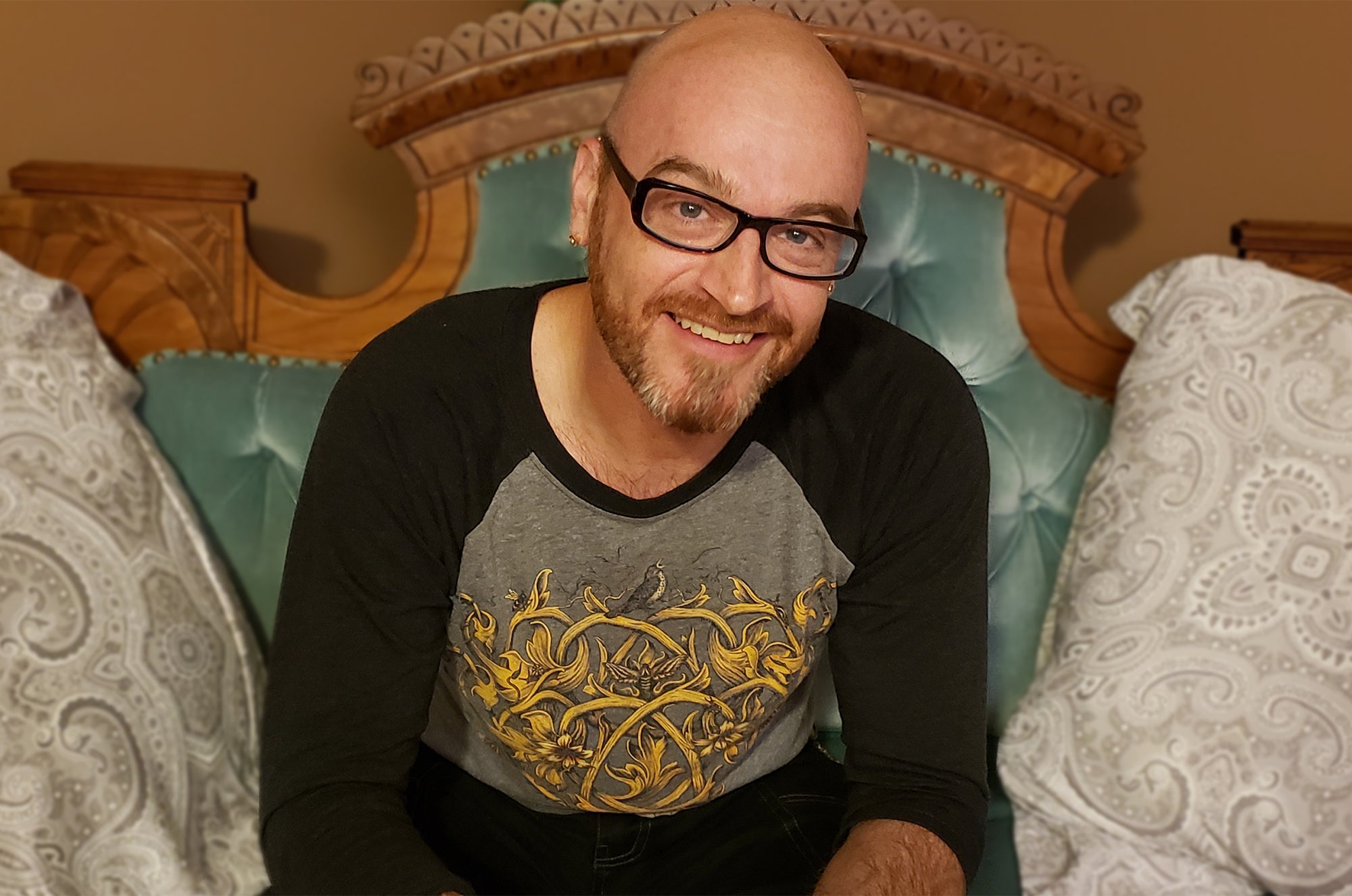 Smiling bald man with glasses and a beard wearing a black and gray shirt with a gold design, sitting on a bed with teal tufted headboard and patterned pillows.