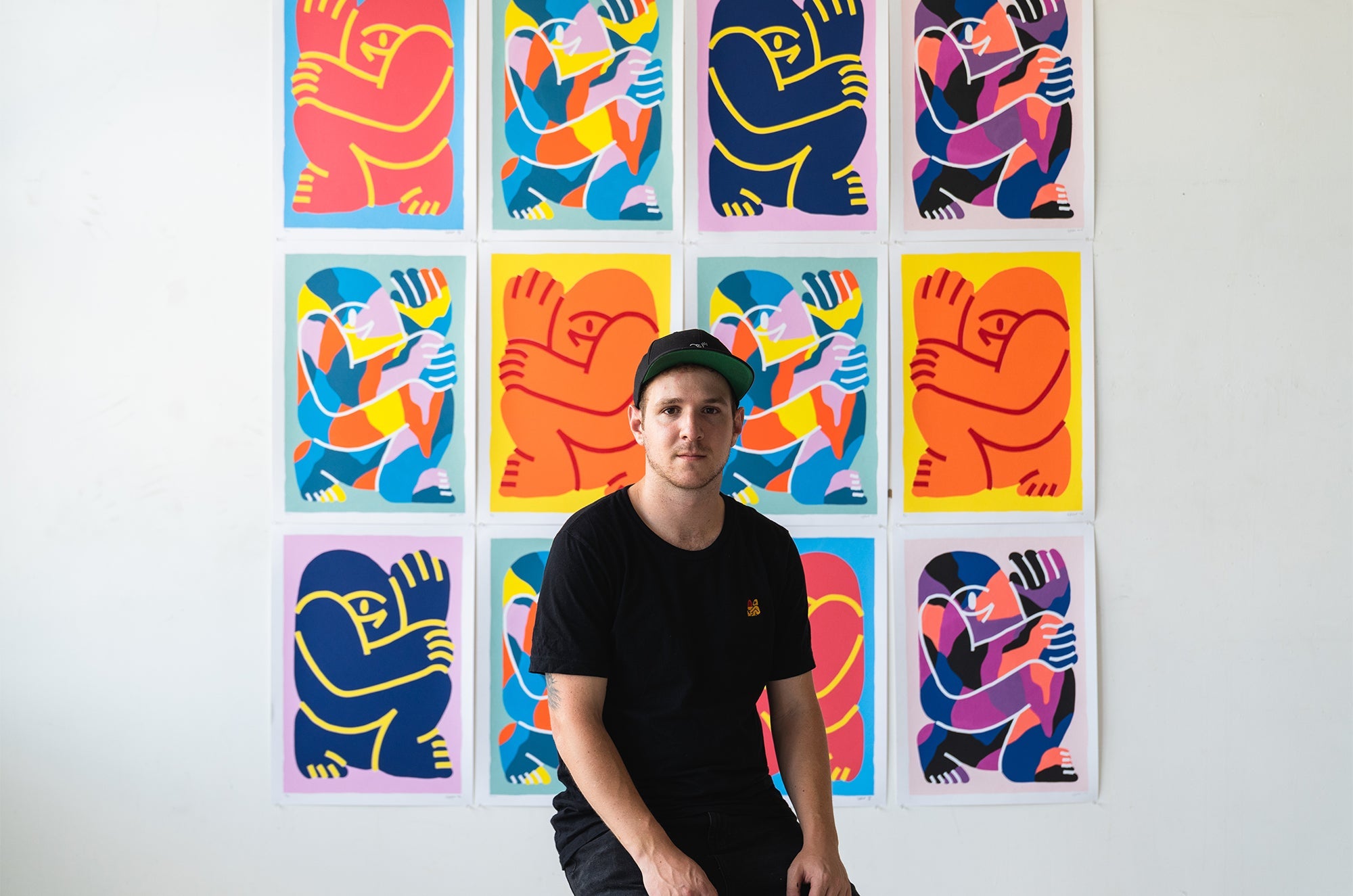 Man in black t-shirt and cap sitting in front of a wall filled with colorful abstract artwork featuring bold, stylized human figures.