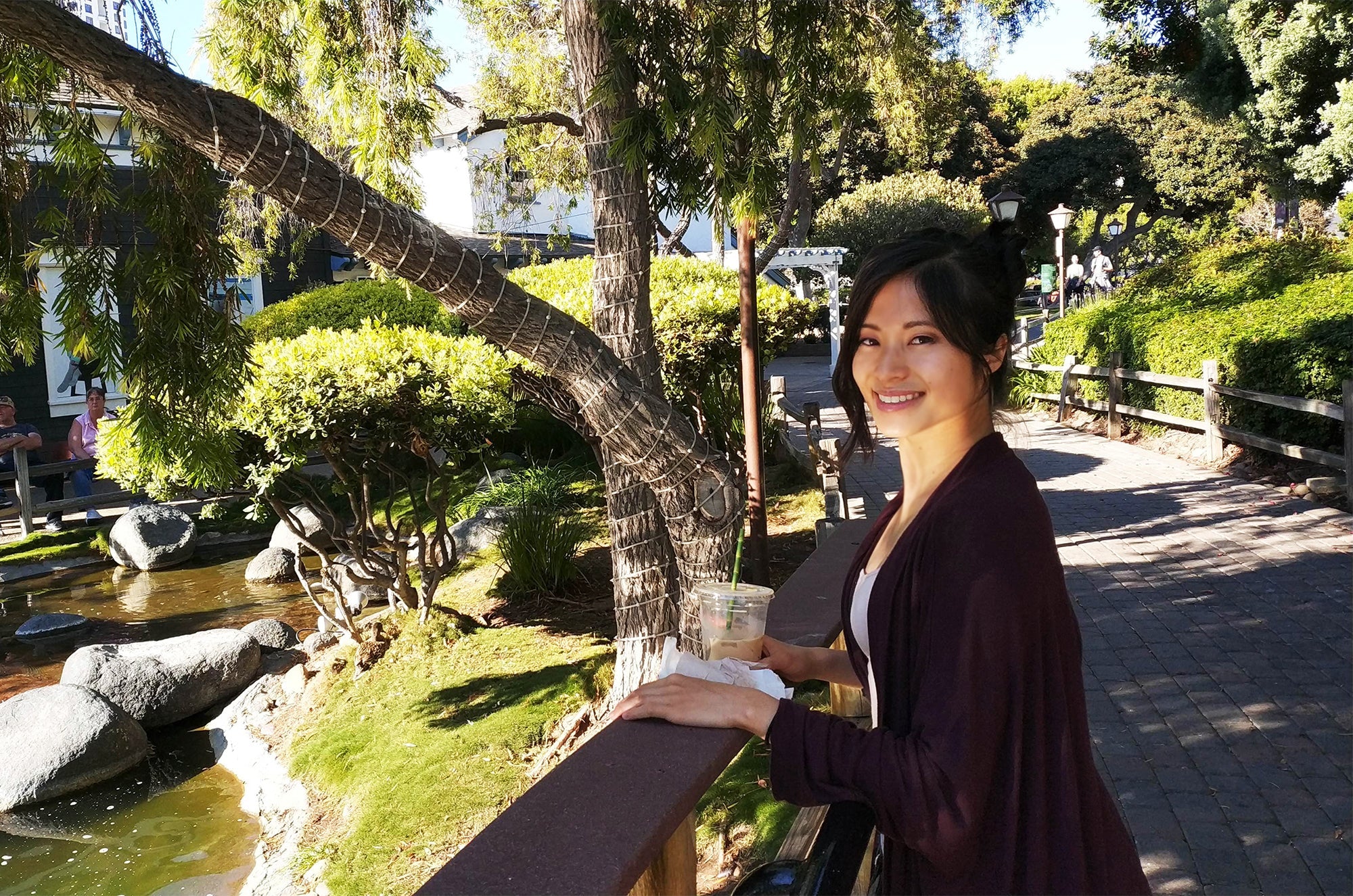 Smiling woman holding a drink while standing near a garden pond with trees, rocks, and sunlight.