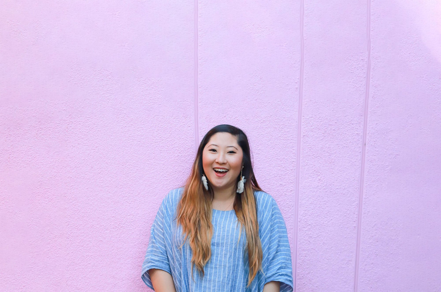 Smiling woman with long ombré hair wearing a blue striped shirt, standing against a bright pink textured wall.