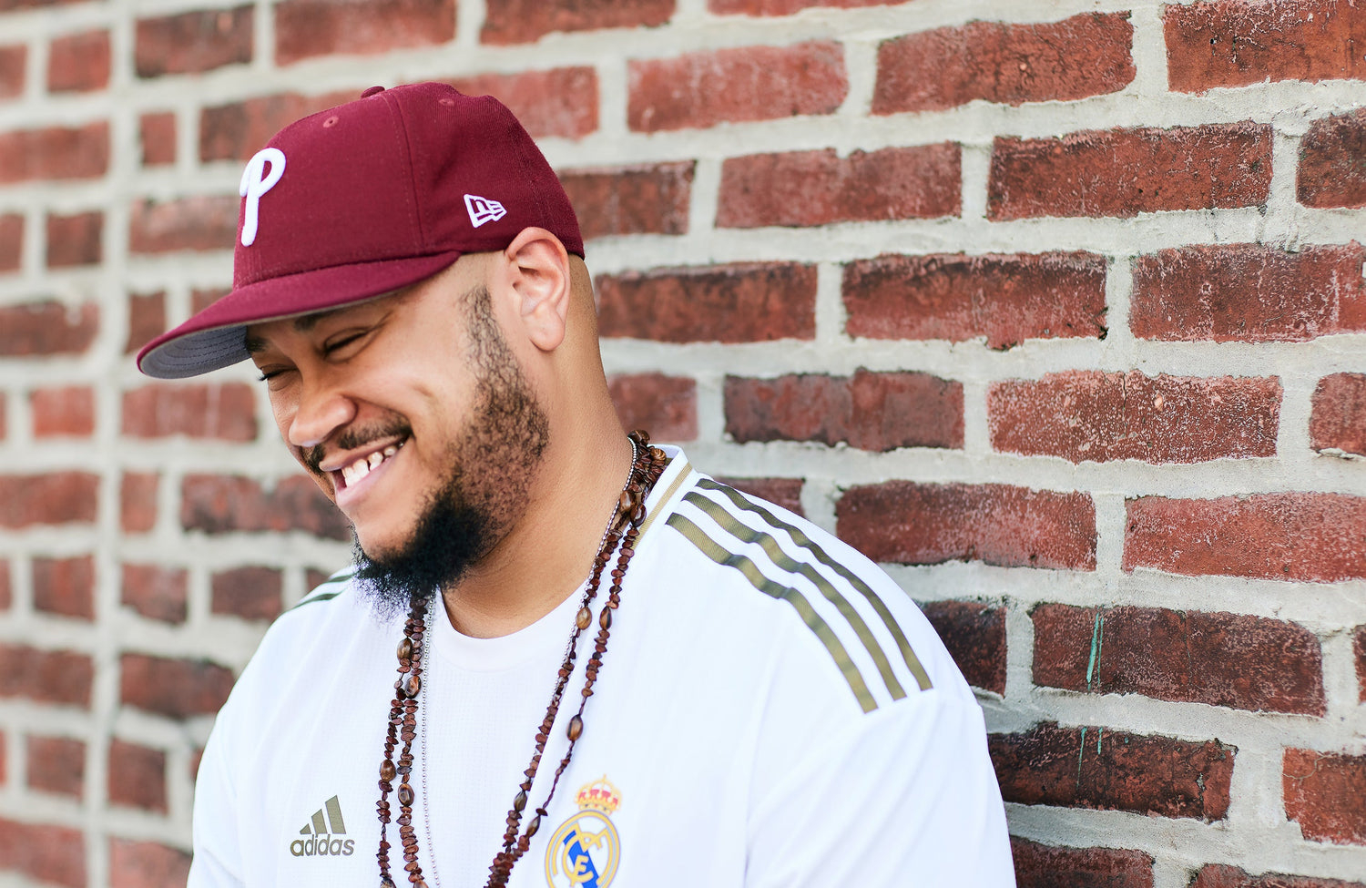 Smiling man wearing a burgundy baseball cap, white sports jersey, and beaded necklace standing in front of a red brick wall.