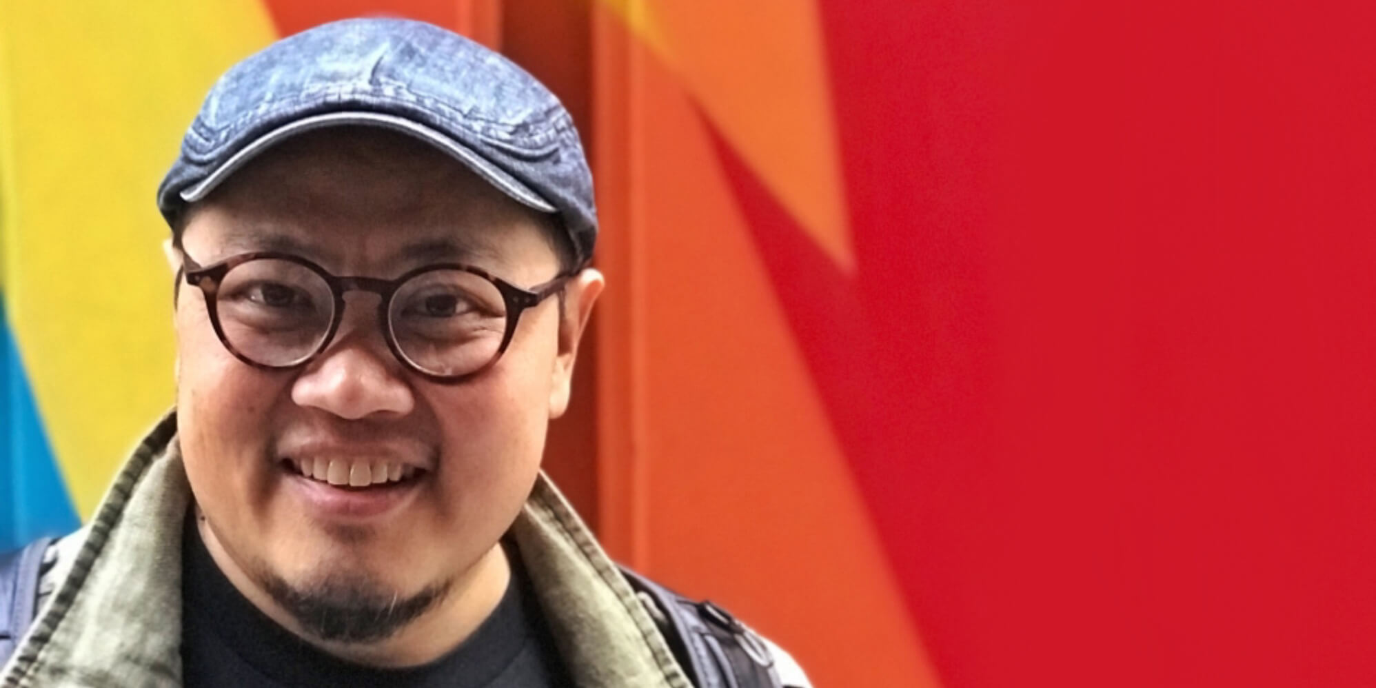 Portrait of a smiling man wearing glasses and a denim cap, standing in front of a colorful red and yellow background.