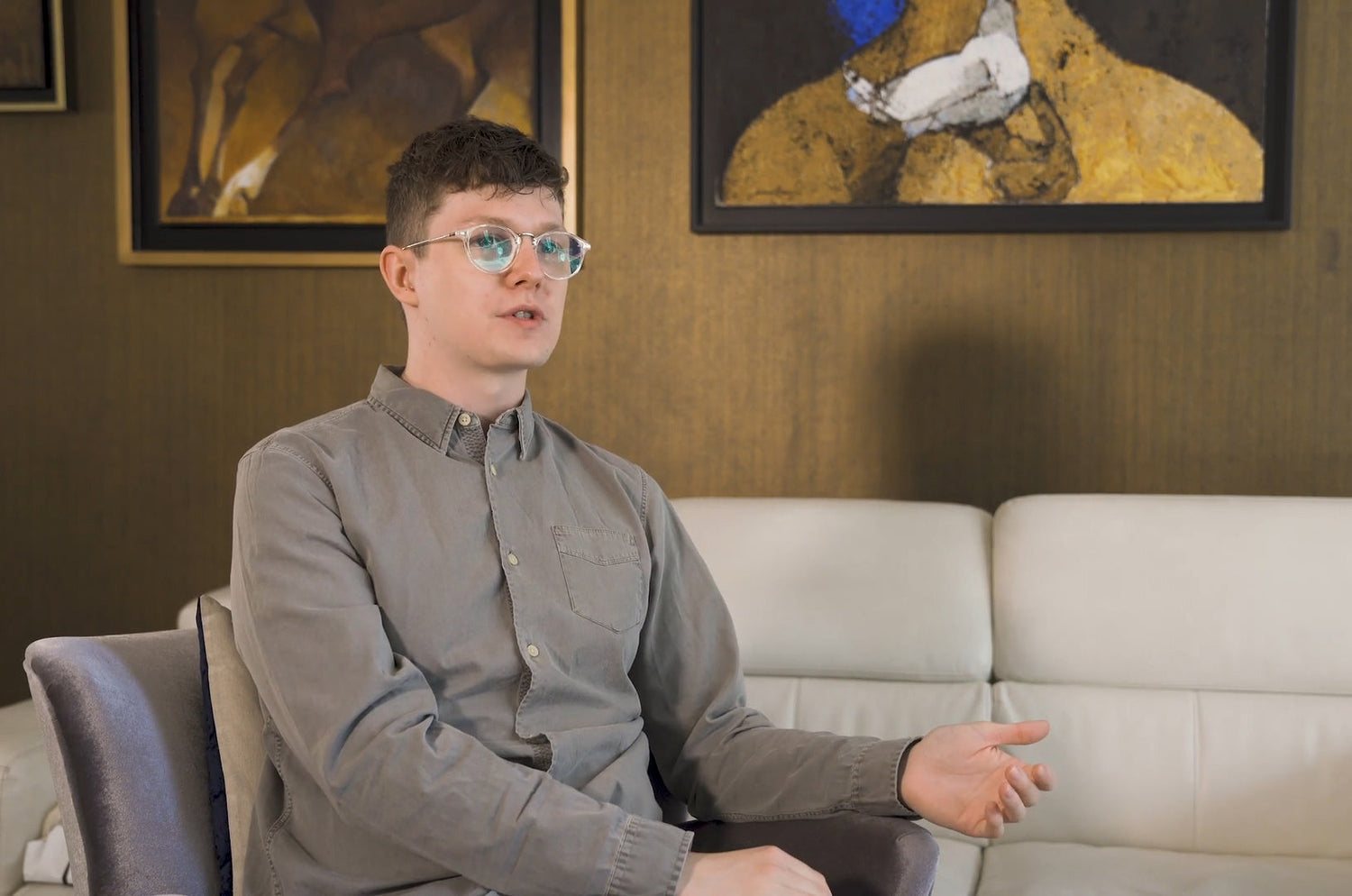 Man wearing glasses and a gray shirt sitting on a sofa, speaking during an interview with abstract paintings in the background.