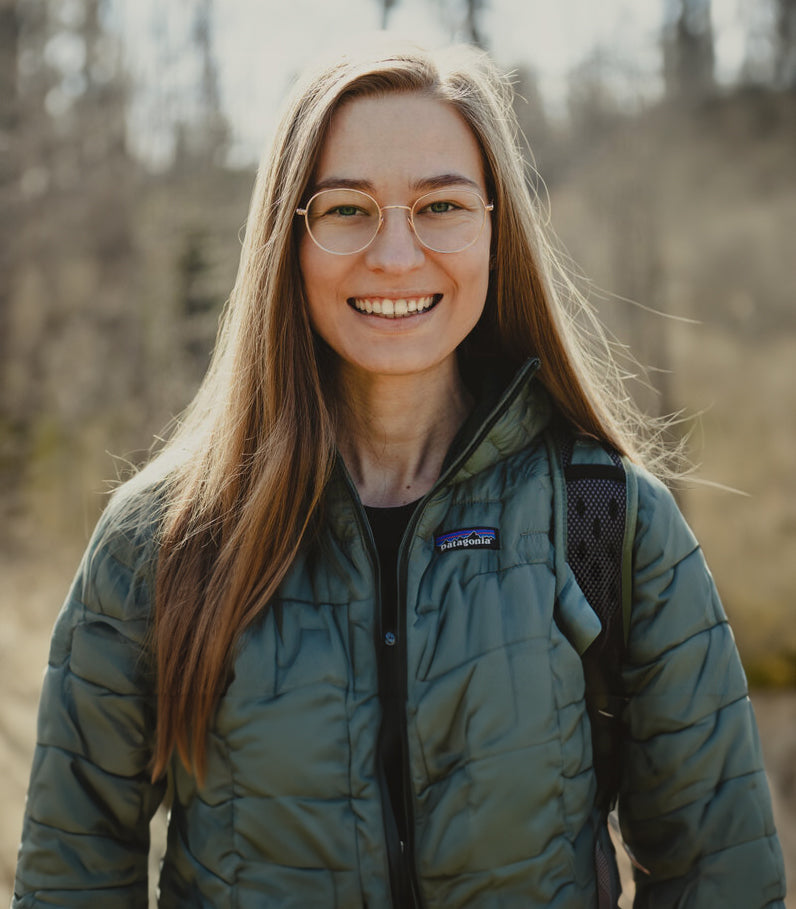 A woman with glasses wearing a fleece jacket and a backpack standing outside and smiling.