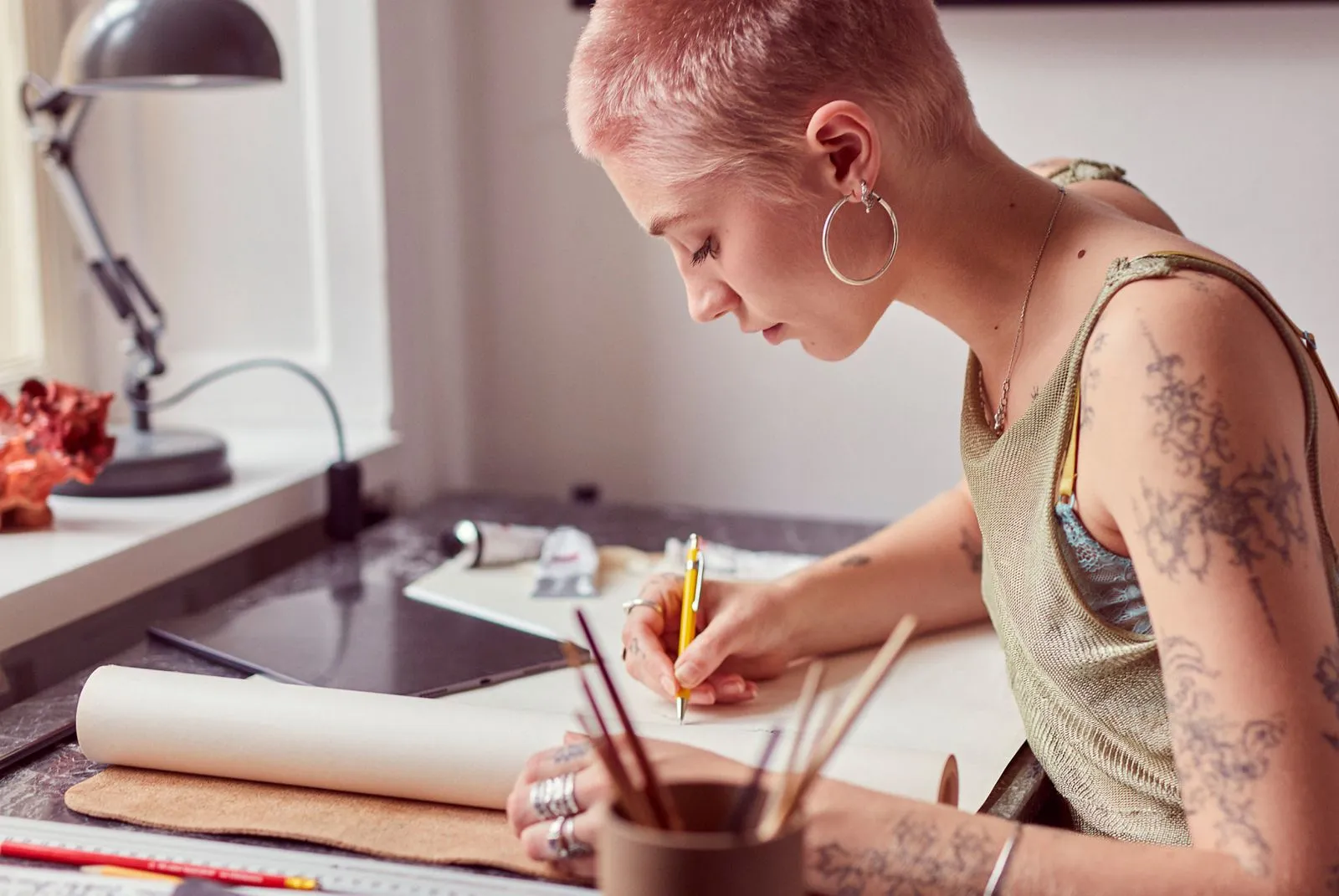 A woman is sketching while sitting at a desk covered with art supplies. An iPad lays atop the desk.