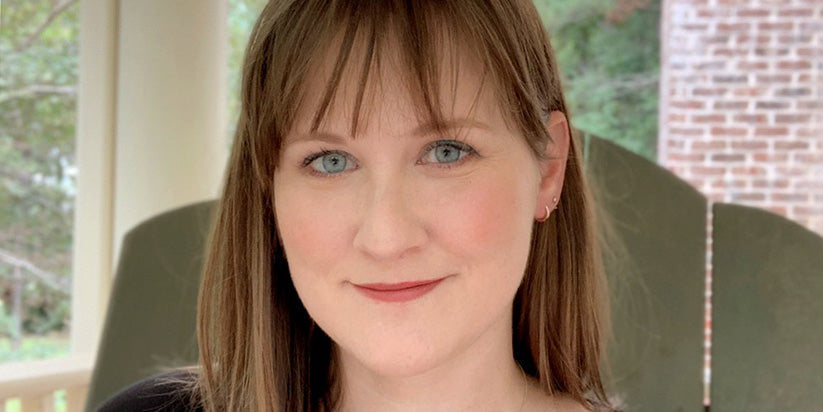 Close-up portrait of a woman with straight light brown hair and bangs, smiling softly while sitting outside on a porch.