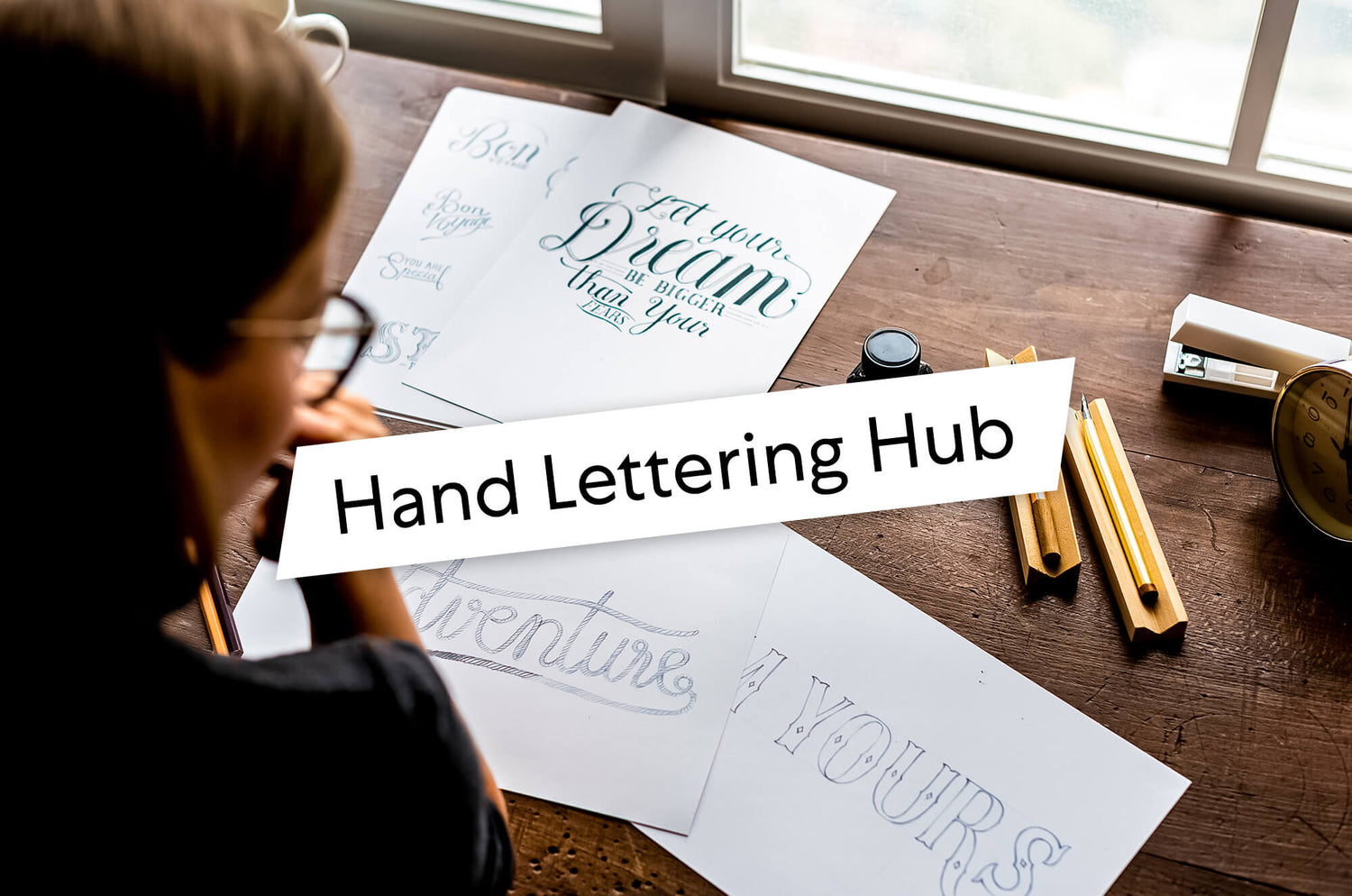 Person working on hand lettering sketches at a wooden desk with pencils, ink, and paper that reads phrases like ‘Let your dream be bigger than your fears.’ Overlay text says: Hand Lettering Hub.
