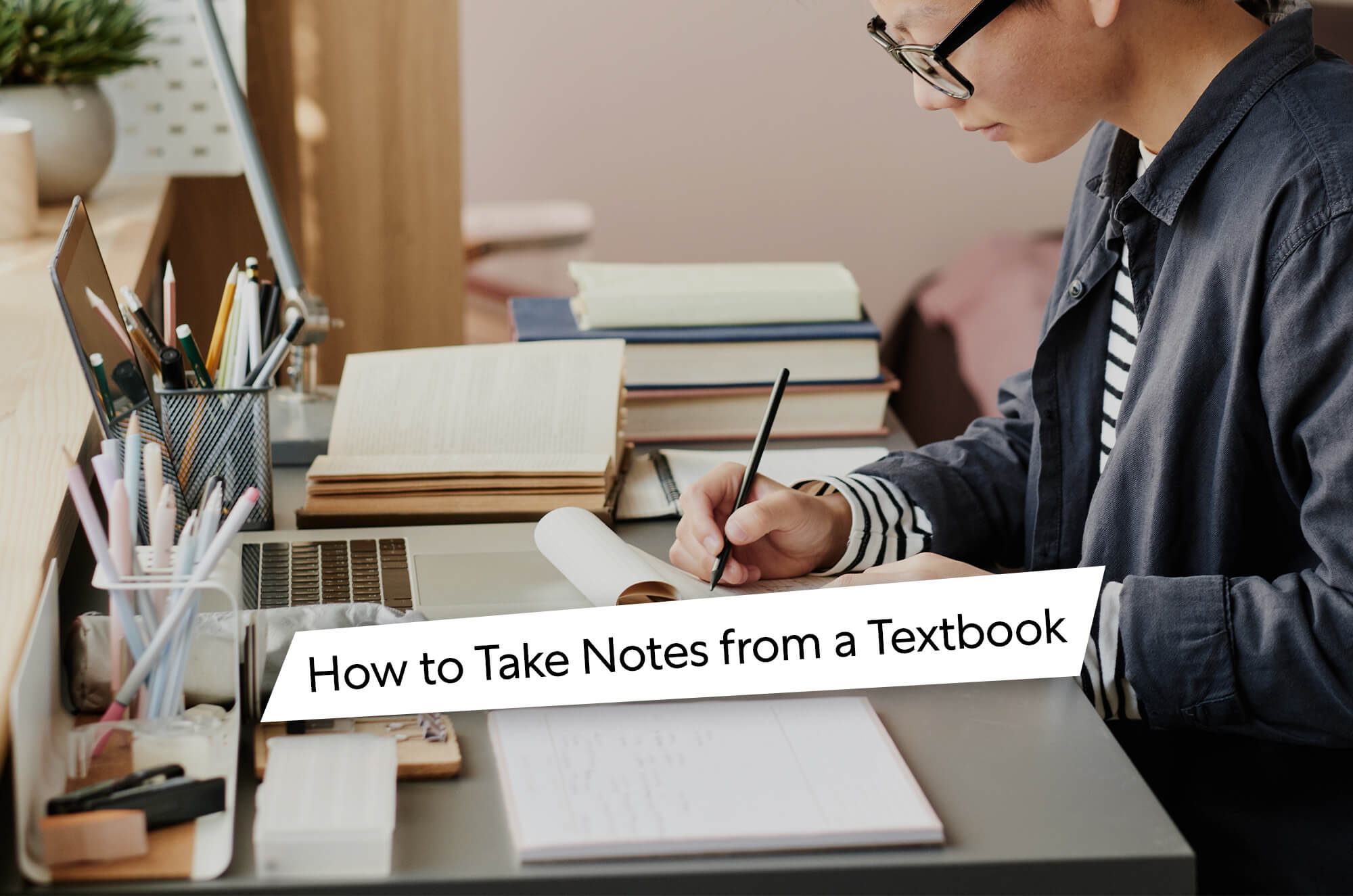 Student writing notes on paper while studying from an open textbook, with stacked books and stationery on the desk.