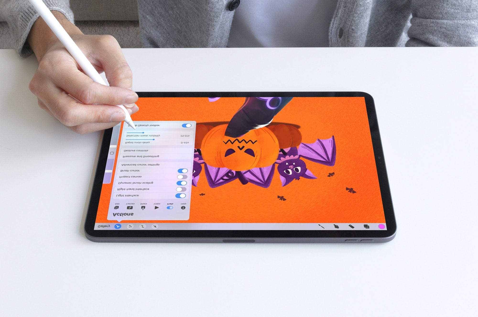 Person using an Apple Pencil on an iPad to draw a Halloween illustration of a pumpkin with bats on an orange background.