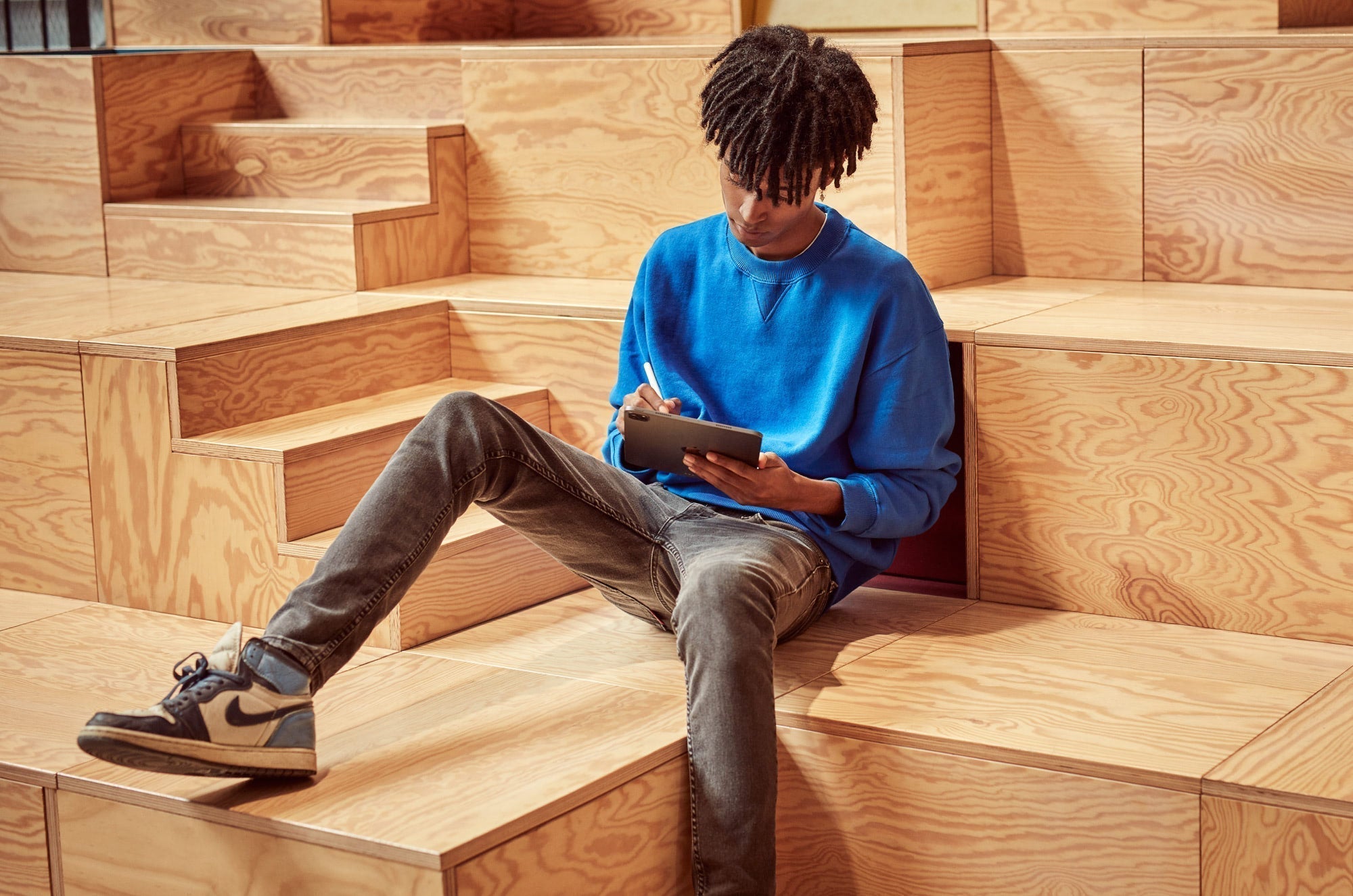 Person in a bright blue sweatshirt sitting on wooden tiered seating, using an iPad with an Apple Pencil.