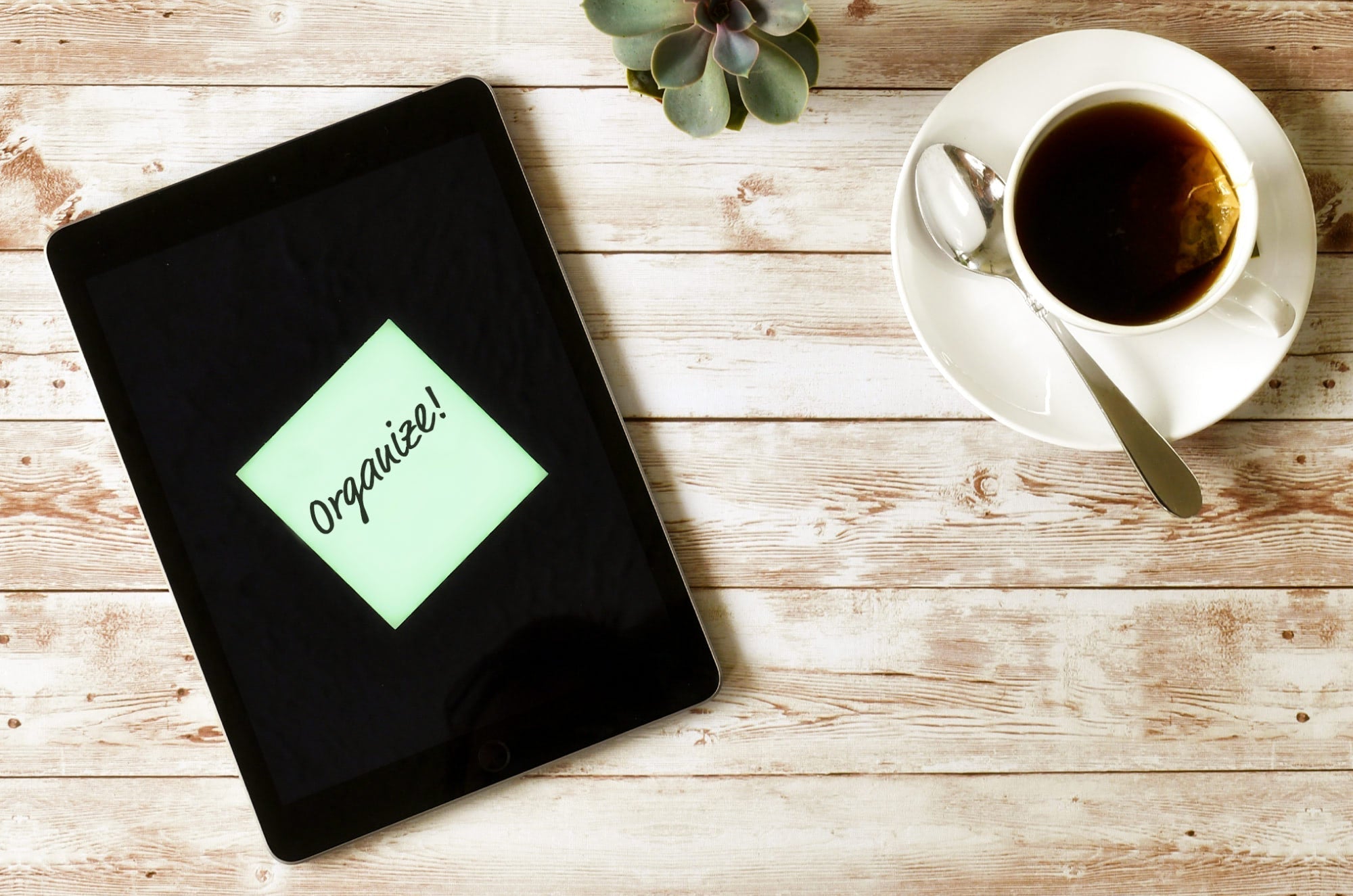 Flat lay of an iPad on a wooden table displaying a digital sticky note with the word ‘Organize!’ next to a cup of tea and a succulent plant.