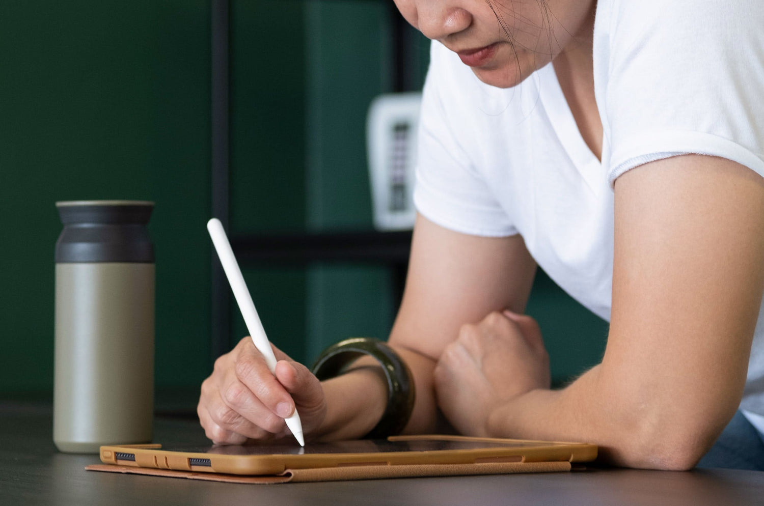 Person in a white t-shirt leaning over a table, using an Apple Pencil on an iPad in a brown case, with a travel mug nearby.