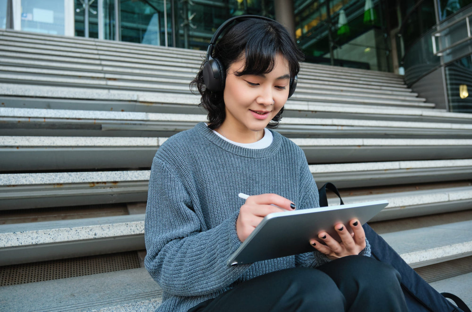 Person wearing headphones and a gray sweater sitting on outdoor steps, smiling while drawing or writing on an iPad with an Apple Pencil.