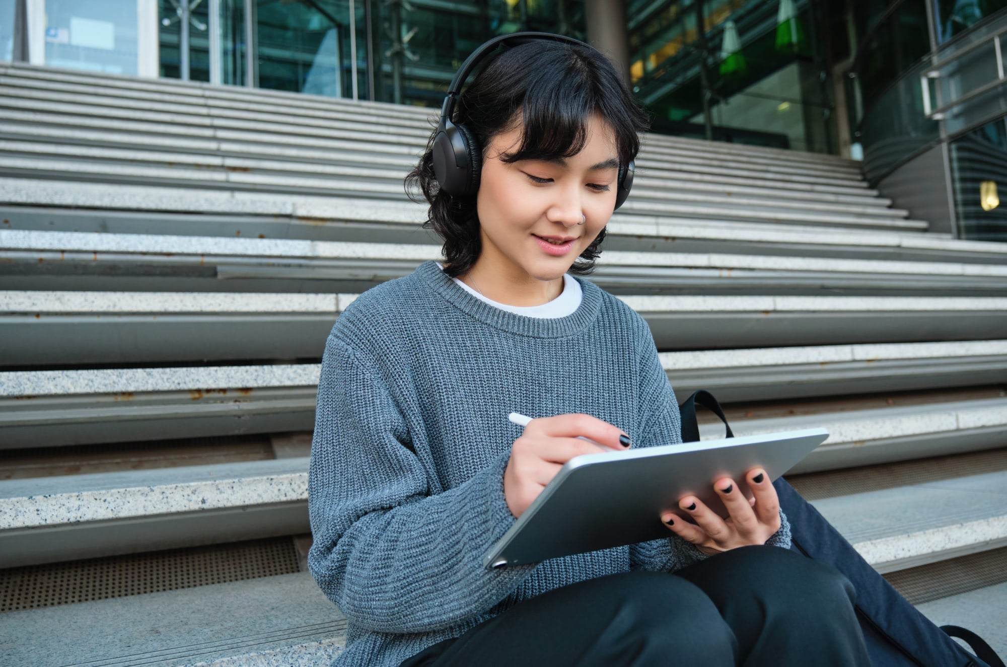 Person wearing headphones and a gray sweater sitting on outdoor steps, smiling while drawing or writing on an iPad with an Apple Pencil.