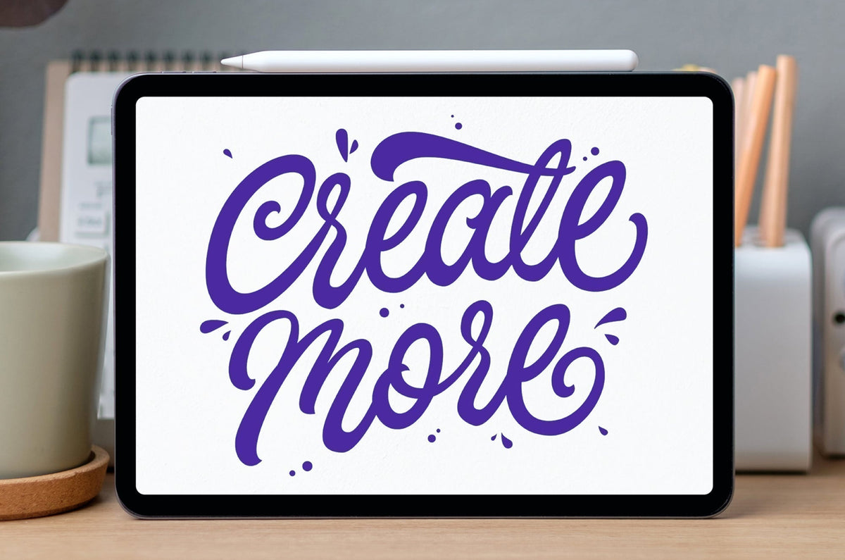 An iPad with Apple Pencil resting on top, displaying purple hand-lettered text that reads ‘Create More.’ The tablet is on a desk with a mug and stationery in the background.
