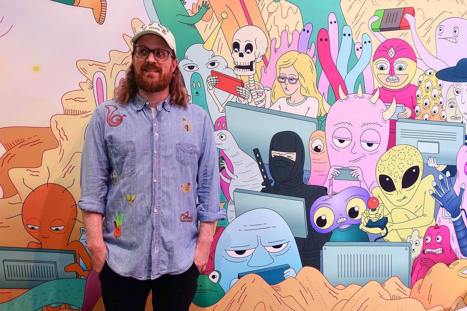 Person in a denim shirt and cap standing in front of a colorful cartoon mural filled with quirky characters, including aliens, skeletons, and ninjas using computers and game controllers.