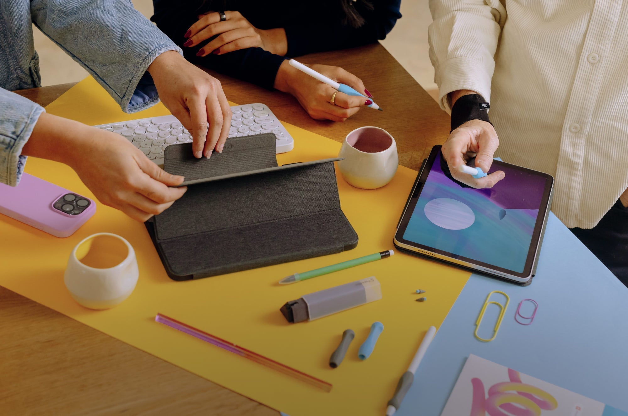 Creative workspace with people using iPads, Apple Pencils, and Paperlike™ accessories on a colorful desk covered with stationery, keyboard, and tablet cases.
