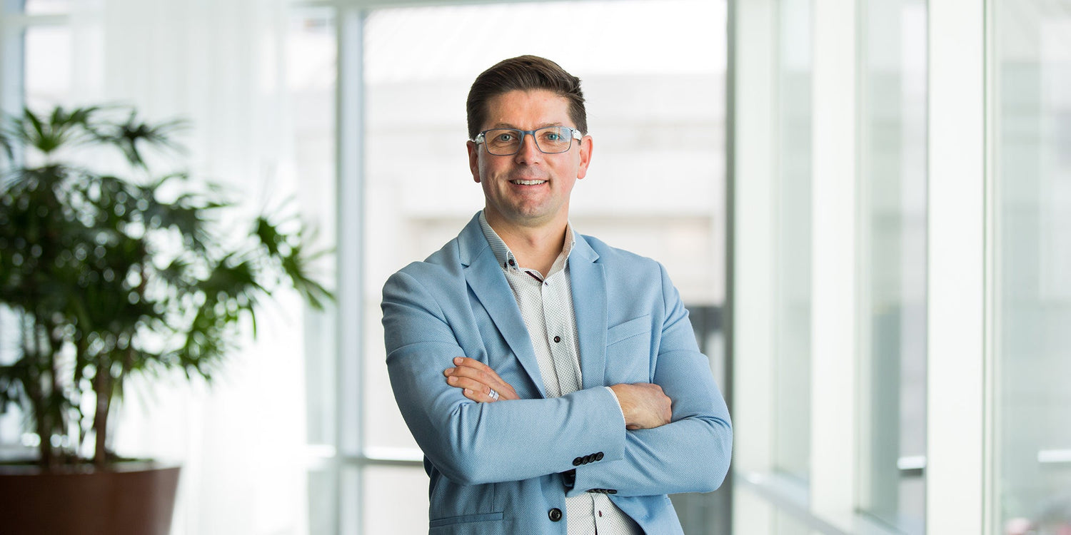 Man in a light blue blazer and glasses standing indoors with arms crossed, smiling at the camera.
