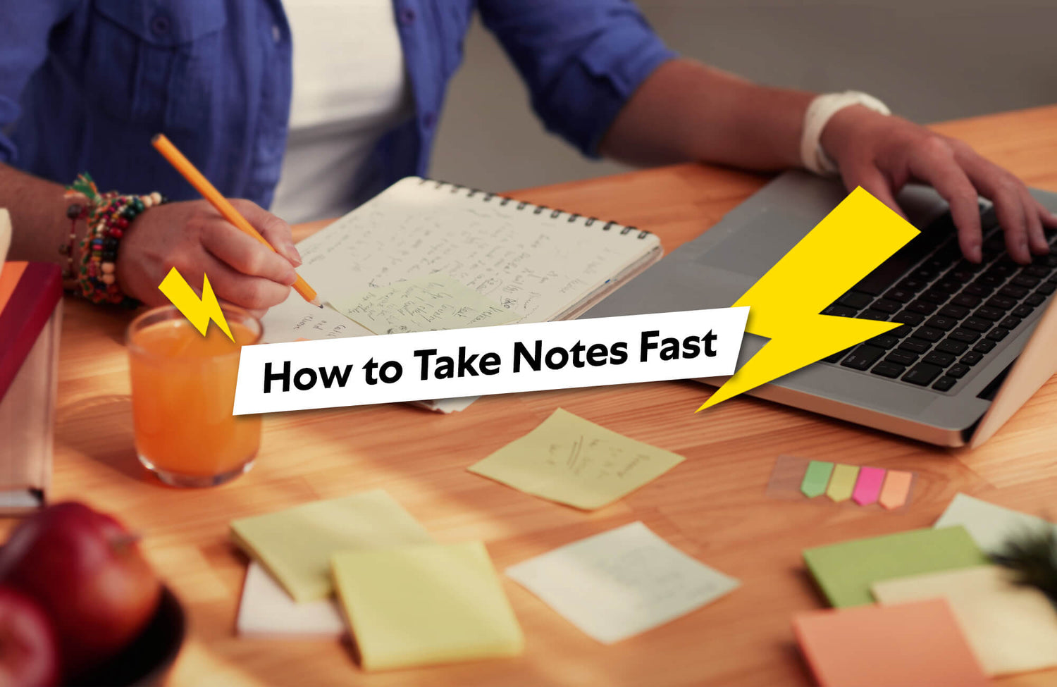 Person taking notes quickly in a notebook while working on a laptop, surrounded by sticky notes and a drink on the desk.