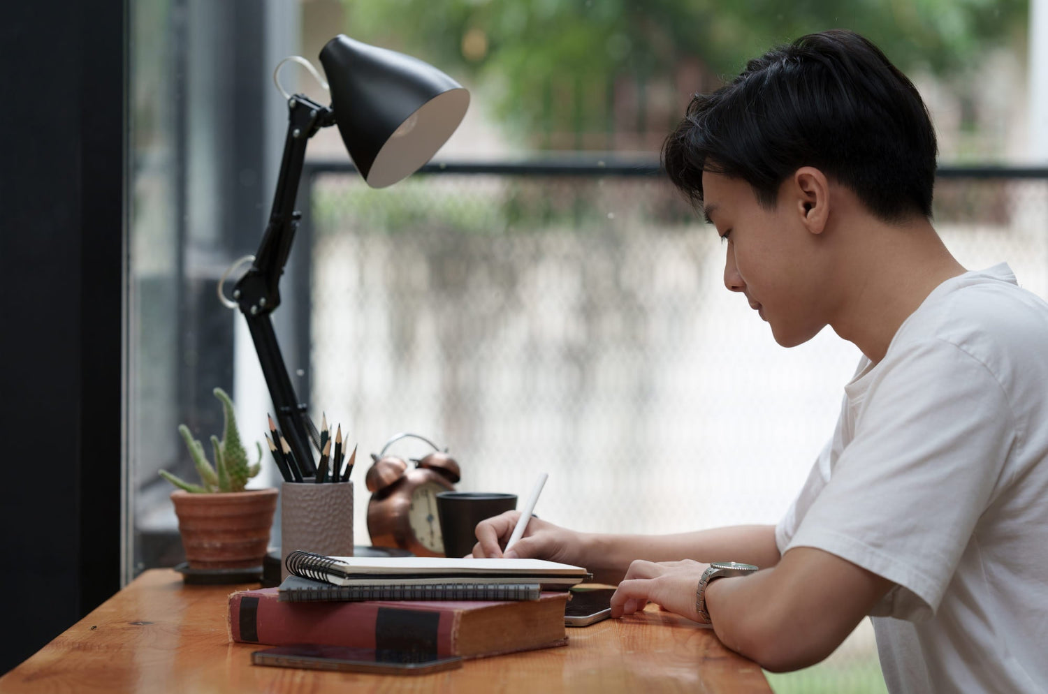 Student sitting at a desk writing in a notebook with pencils, books, and a lamp nearby.