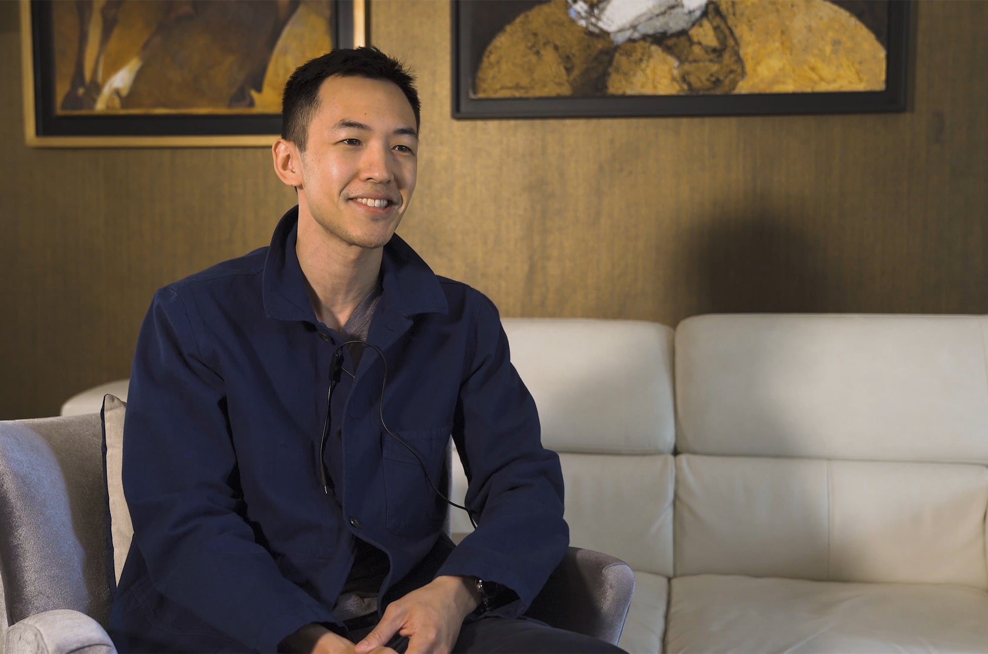 Person seated on a sofa in a warmly lit room, wearing a navy jacket and smiling during an interview.