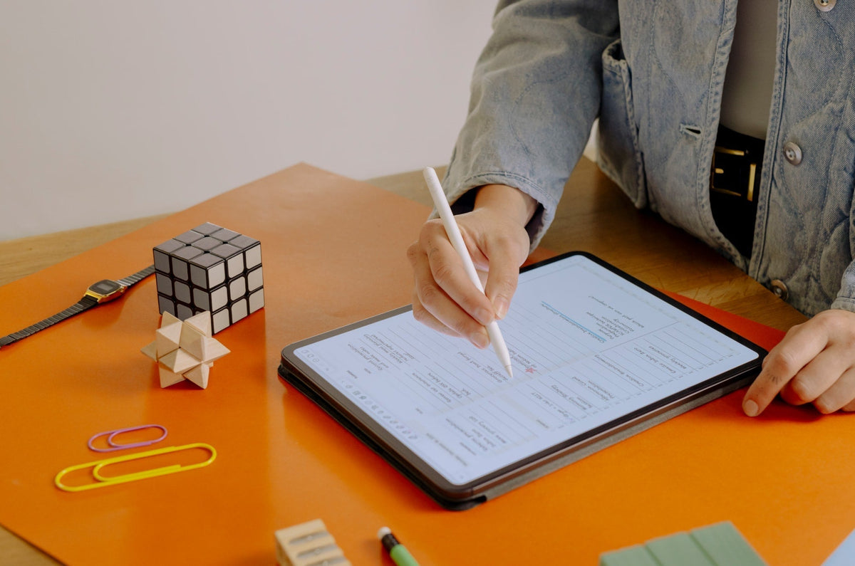 Person using an Apple Pencil on an iPad to fill out a digital planner, with office supplies, a Rubik’s cube, and a wooden puzzle on an orange desk mat.