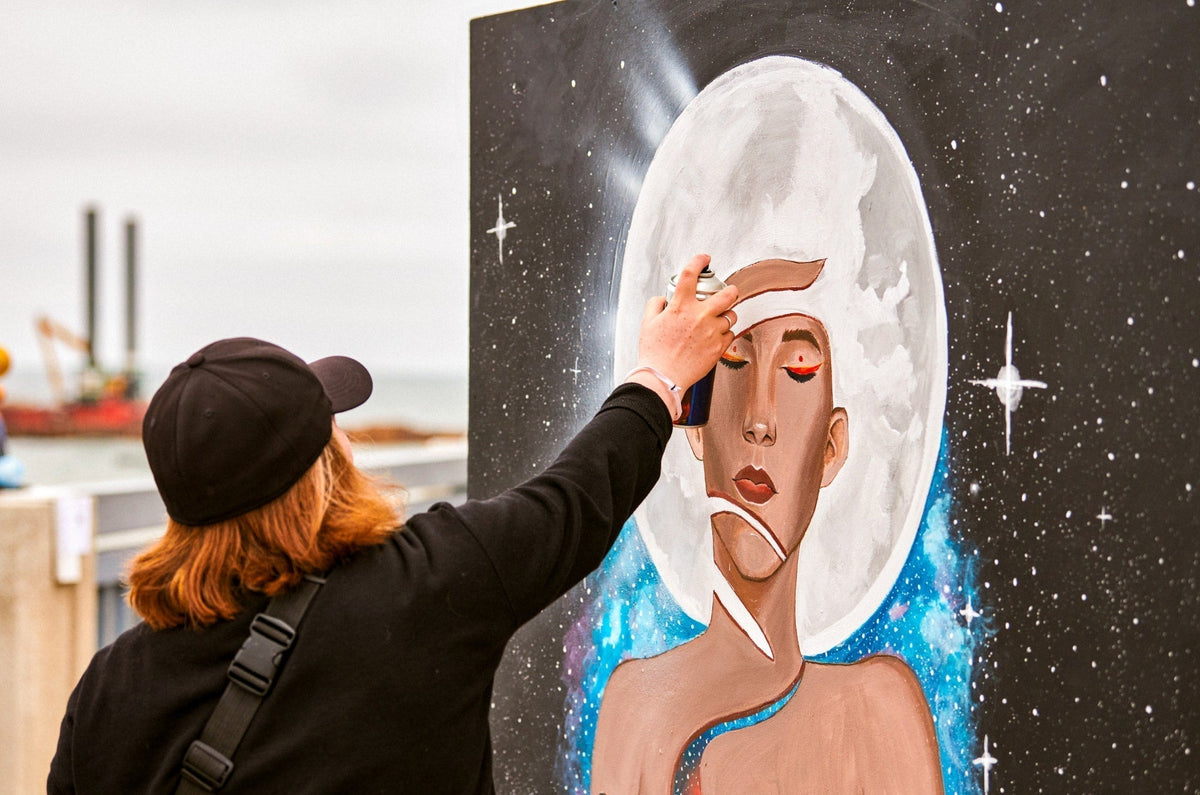 Female artist in black cap is painting picture with paint spray can spraying it onto canvas at outdoor street exhibition, side view of female art maker.