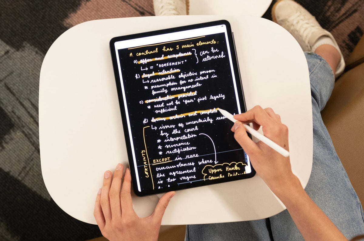 Person taking handwritten digital notes on an iPad with an Apple Pencil. The screen shows neatly written text about contract law on a black background with yellow highlights. The iPad rests on a small white table.