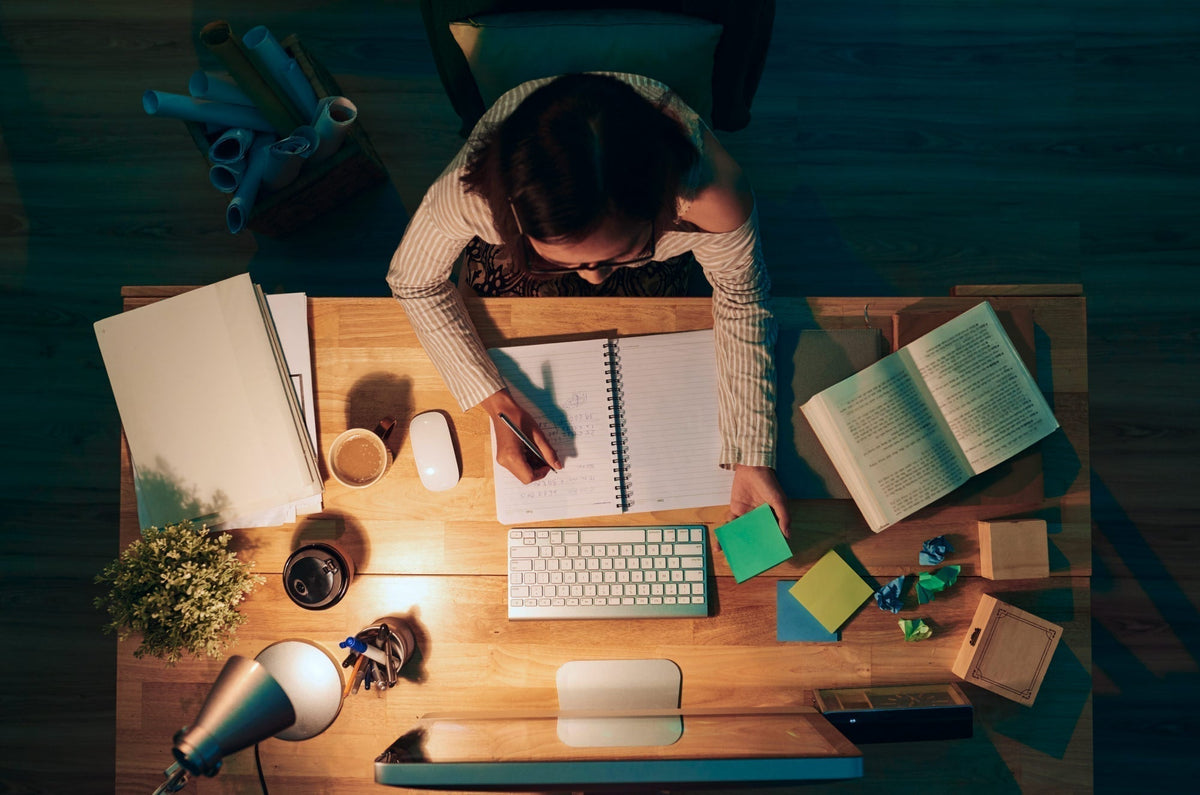 An overhead view of a woman sitting at a desk, writing in a notebook, with books and other accessories on the desk.