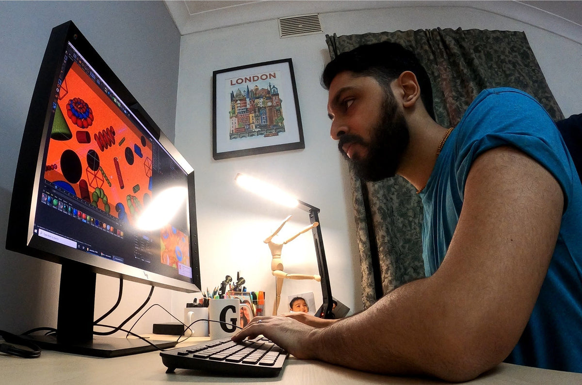 Man working at a desktop computer with colorful 3D artwork displayed on the screen, seated at a desk with lamp, pens, and framed London artwork on the wall.