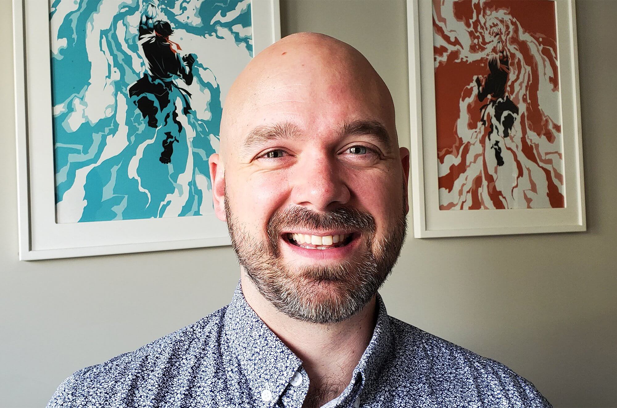 Smiling bald man with a beard wearing a patterned button-up shirt, standing indoors with two colorful abstract art prints on the wall behind him.