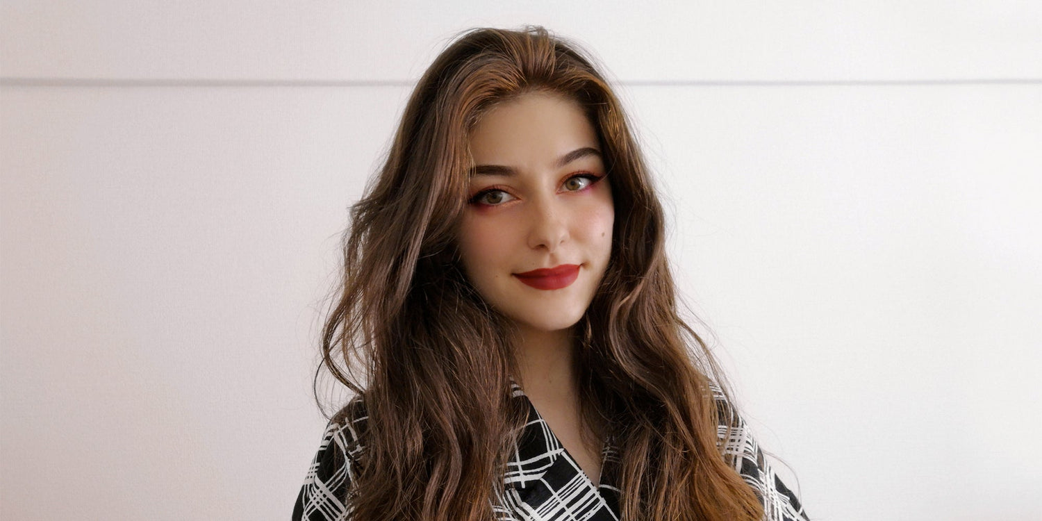Portrait of a young woman with long wavy brown hair, wearing a black and white patterned top, smiling softly in front of a plain white background.