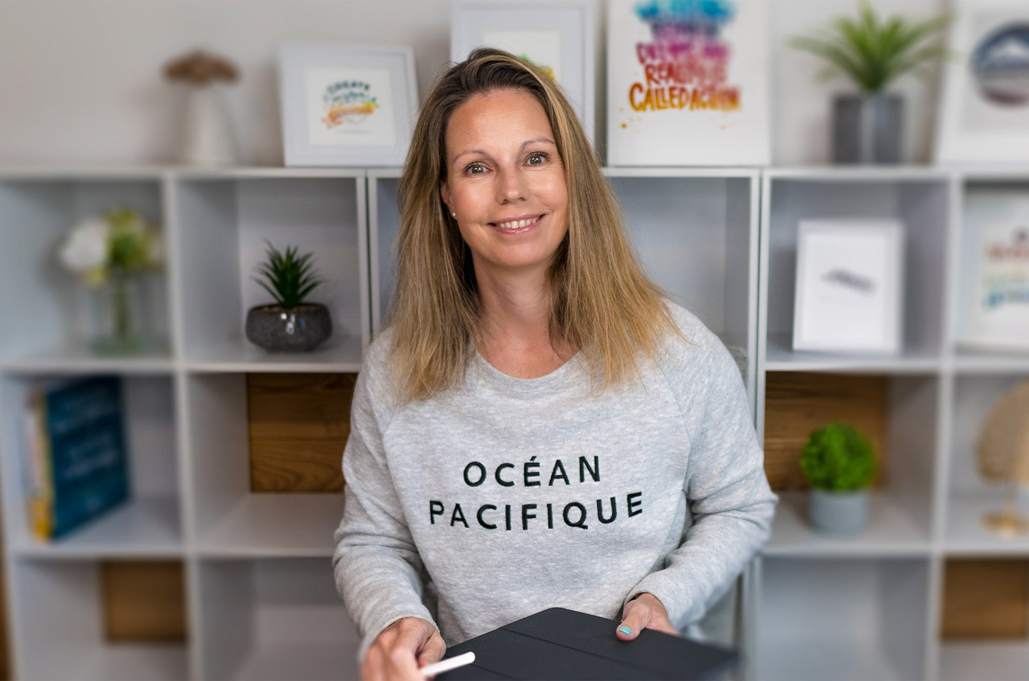 Smiling woman holding an iPad and Apple Pencil, wearing a gray sweater that reads ‘Océan Pacifique,’ seated in front of a white shelving unit with framed art and plants.