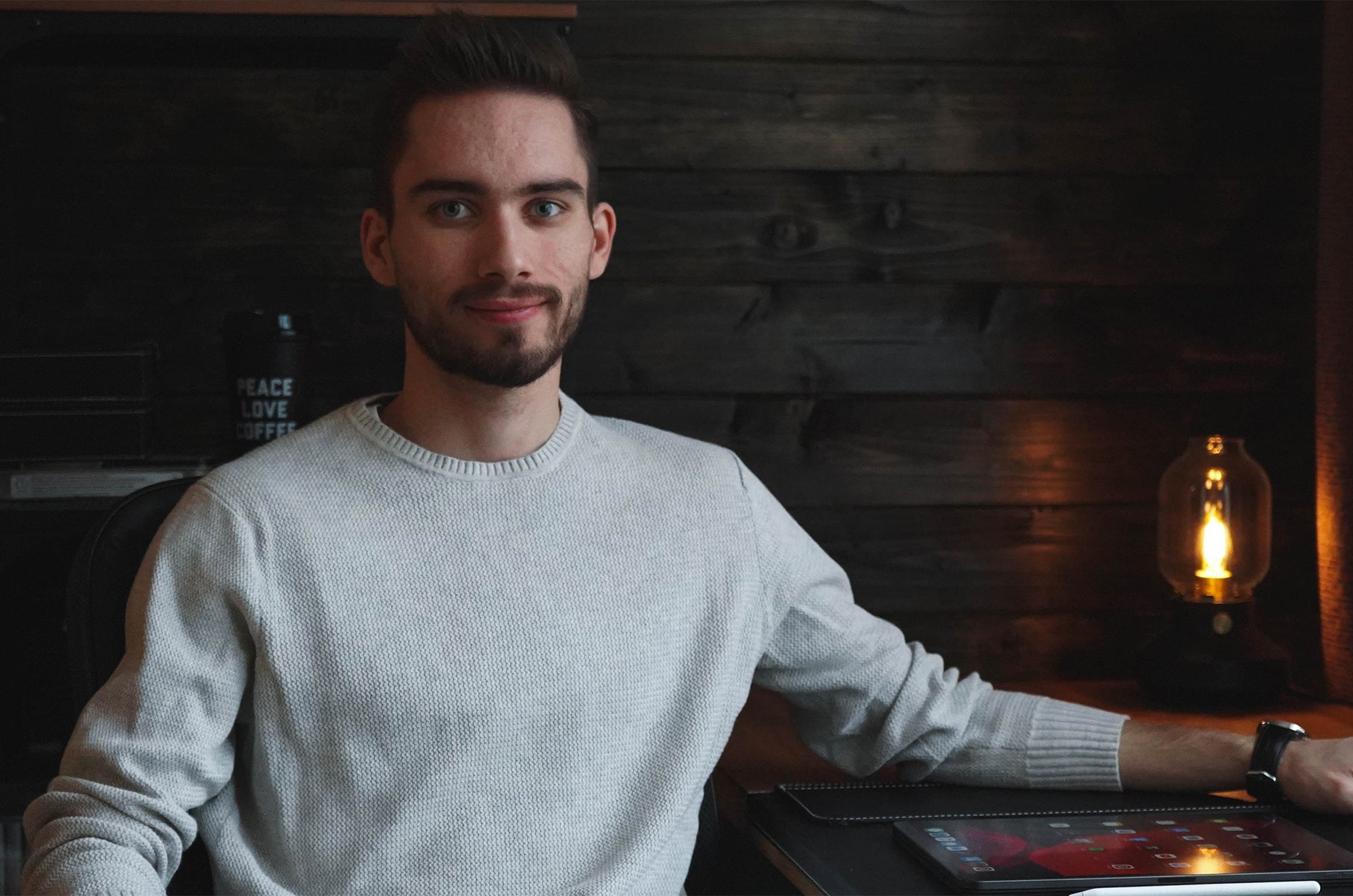 Portrait of a man in a light gray sweater sitting at a desk with a tablet, dark wooden wall and warm lamp in the background.