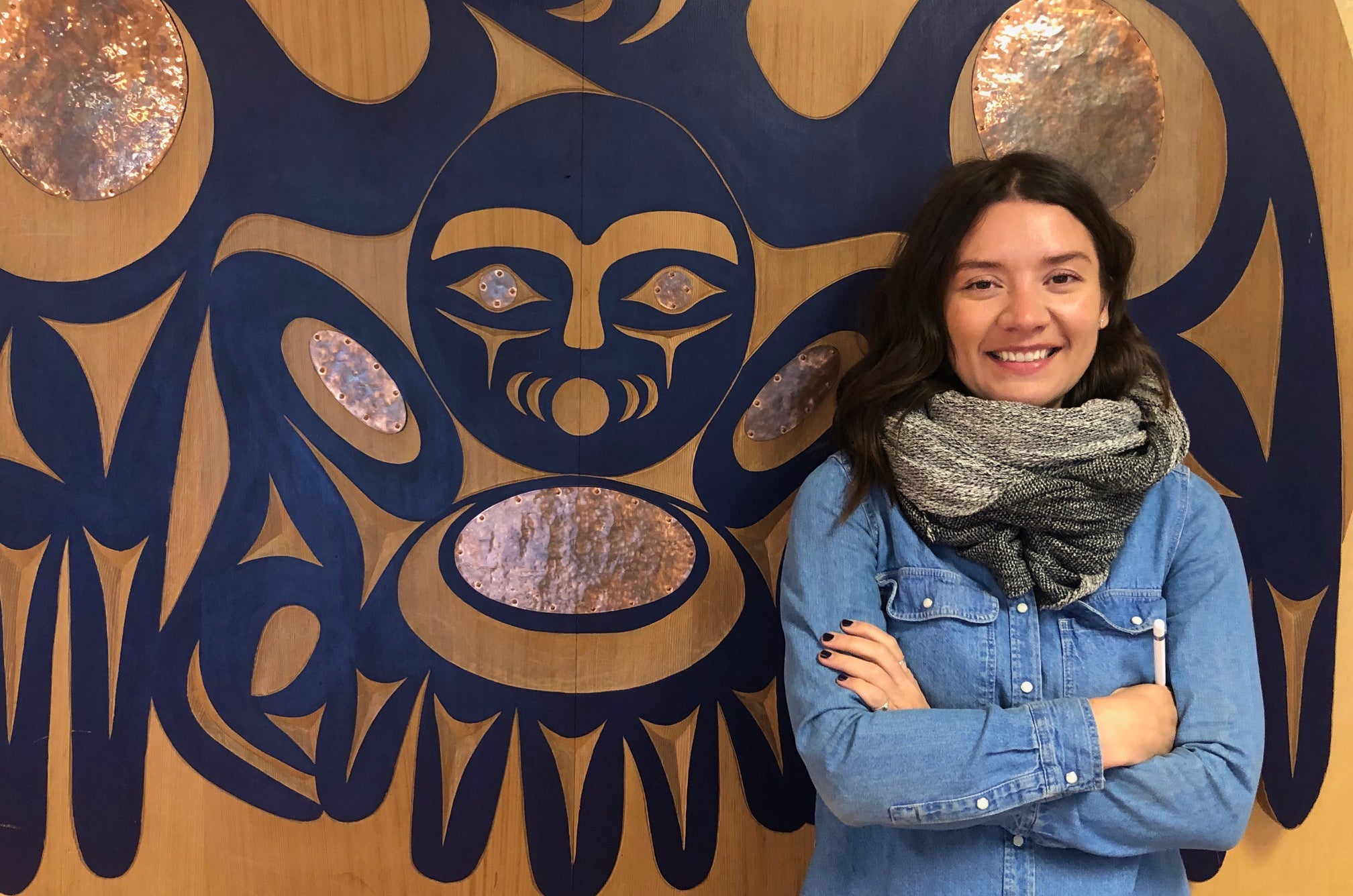 Smiling woman with long dark hair, wearing a denim shirt and gray scarf, standing with arms crossed in front of a large blue and copper Indigenous-style artwork.