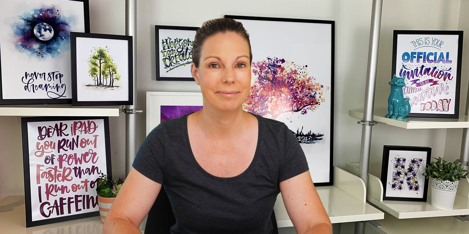 Woman sitting in a studio surrounded by framed calligraphy and watercolor artwork, smiling at the camera.