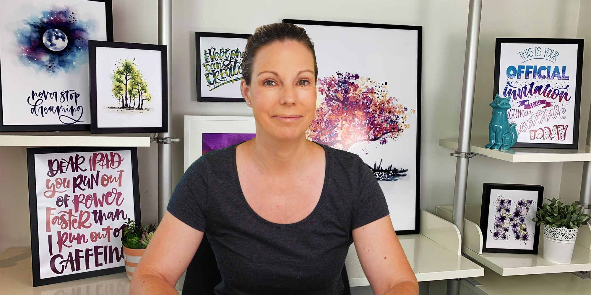 Woman sitting in a studio surrounded by framed calligraphy and watercolor artwork, smiling at the camera.